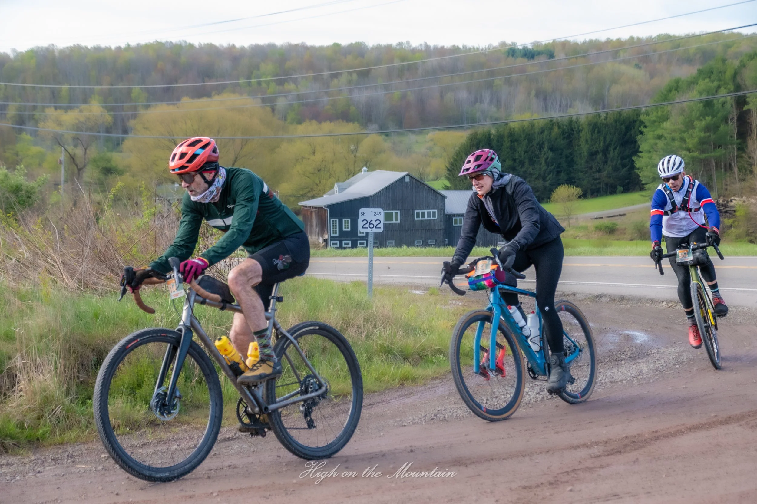 Three cyclists riding on a dirt path beside a rural road with a black barn and green hills in the background. The sky is overcast.