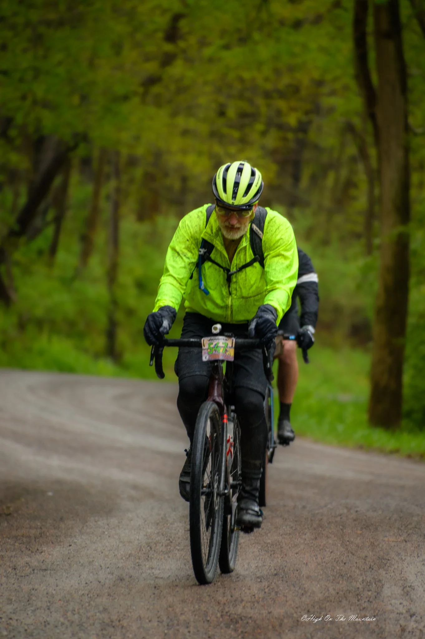 A man wearing a yellow high-visibility jacket and a bicycle helmet riding a mountain bike on a trail through a forest.