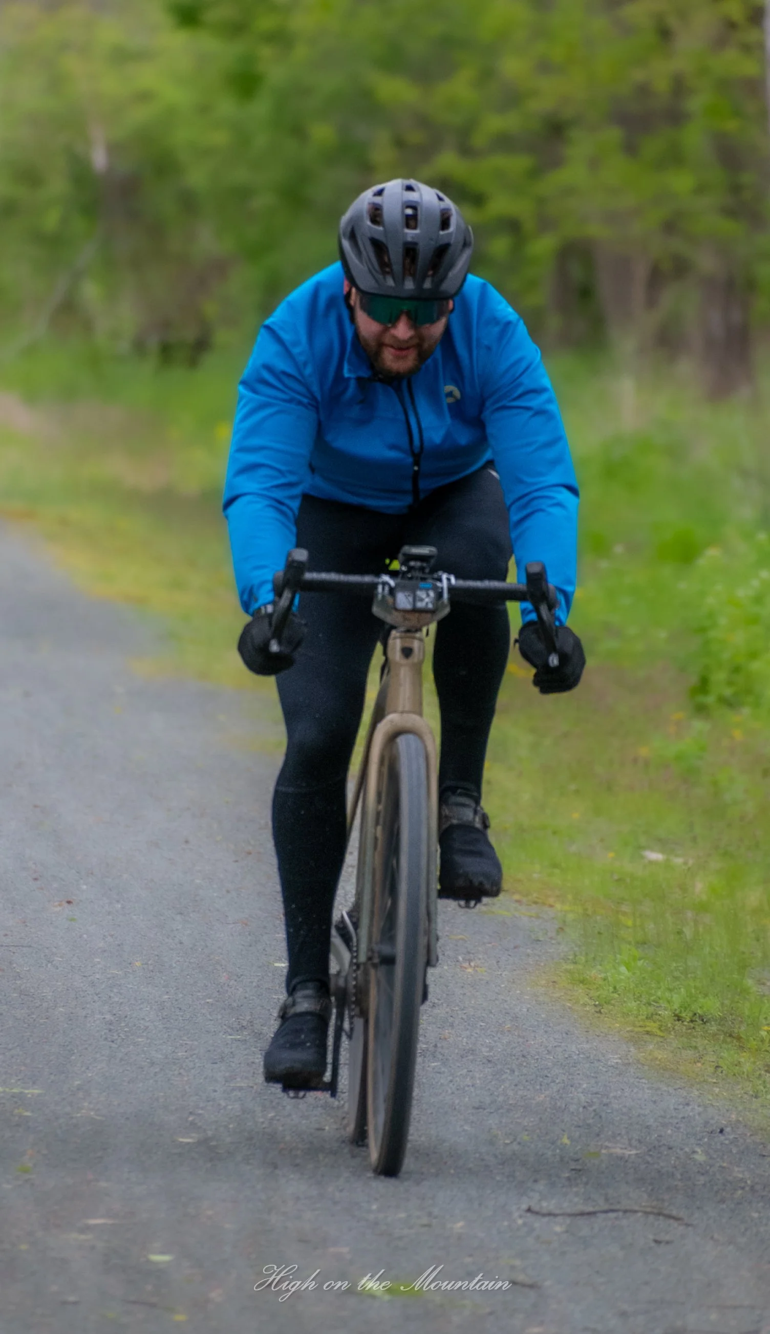 A man wearing a black helmet, sunglasses, a blue jacket, and black gloves riding a mountain bike on a forest trail with green trees in the background.