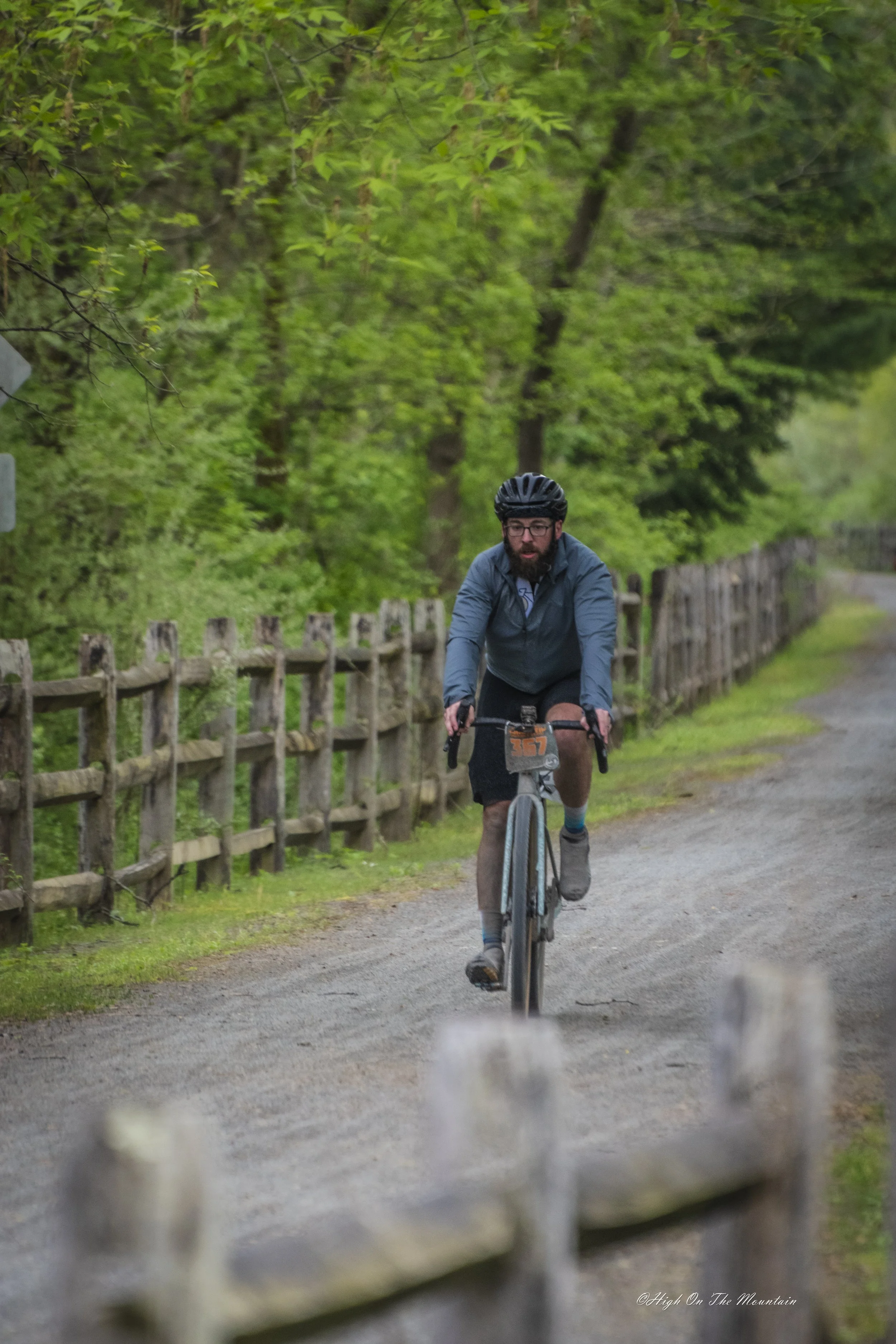 A man with a beard and glasses riding a bicycle on a dirt path surrounded by green trees, with a wooden fence along the side of the trail.