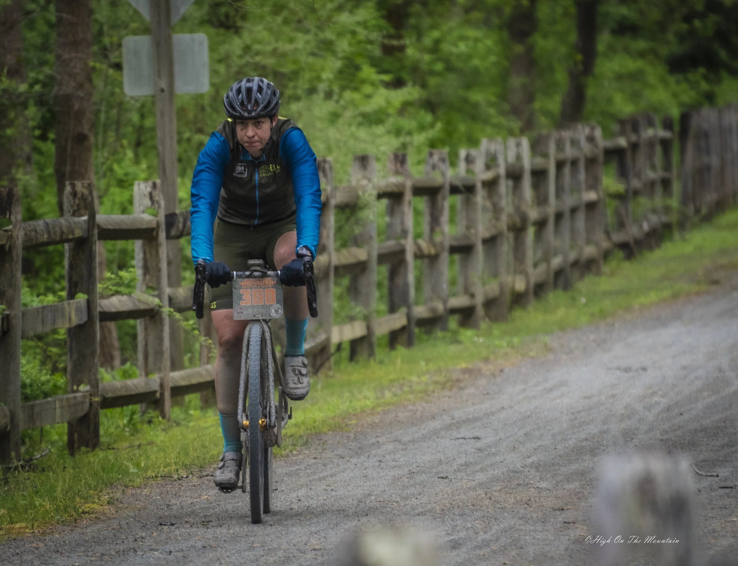 A man riding a mountain bike on a dirt trail through a green forest, wearing a black helmet, blue jacket, and black vest with the number 300 attached to the front of the bike.