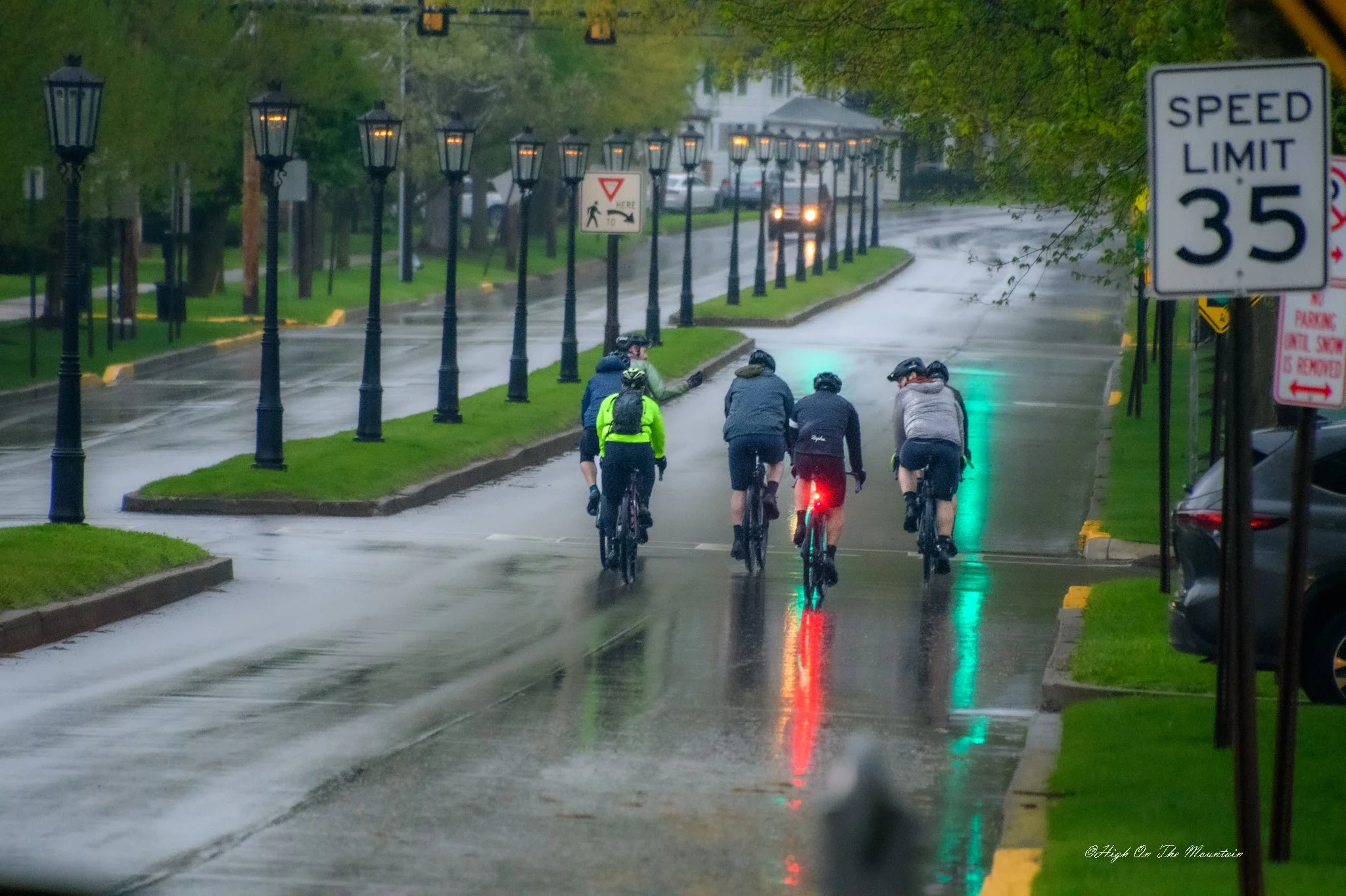A group of cyclists riding on a wet city street during rainy weather at dusk or dawn, with streetlights illuminating the scene.