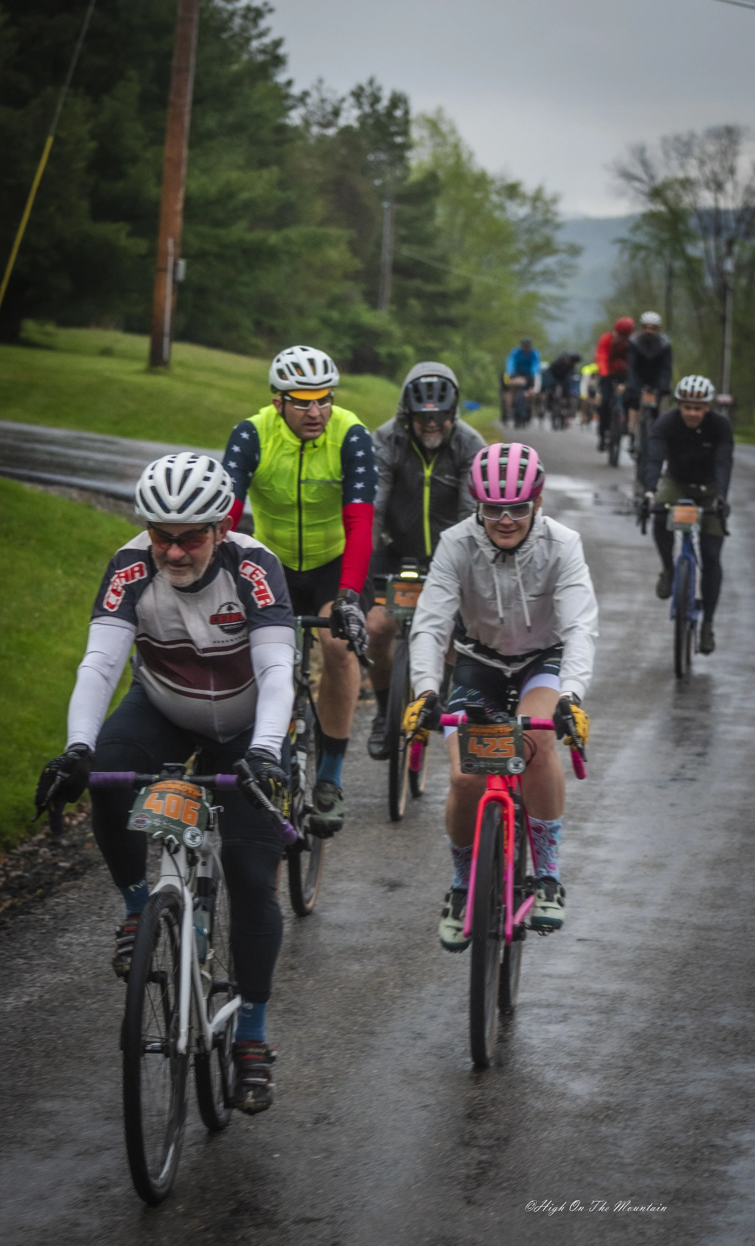 Group of cyclists riding on a wet, paved road surrounded by green trees in a rural area