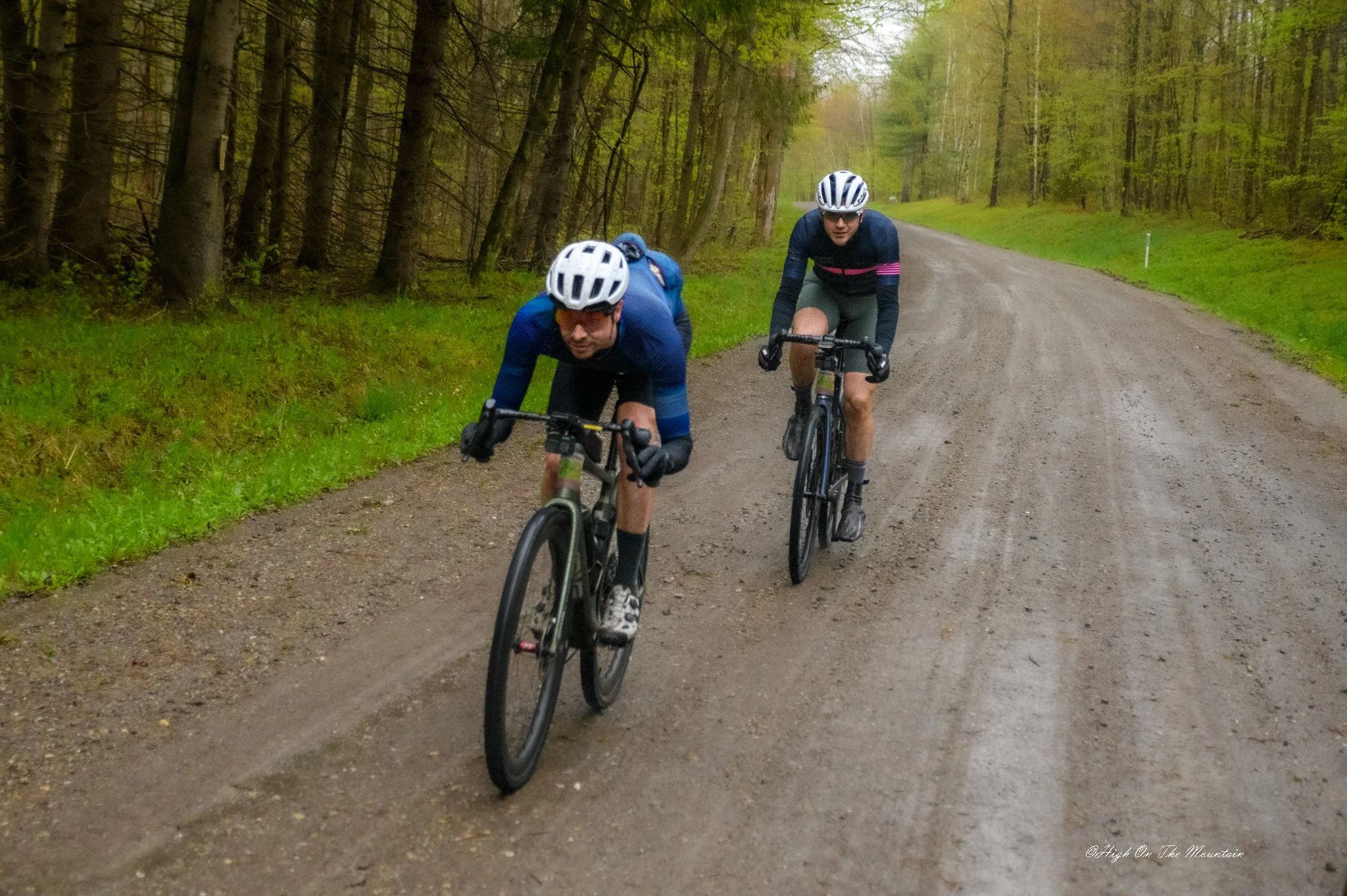 Two cyclists riding on a gravel path through a green forest, wearing helmets and sportswear.