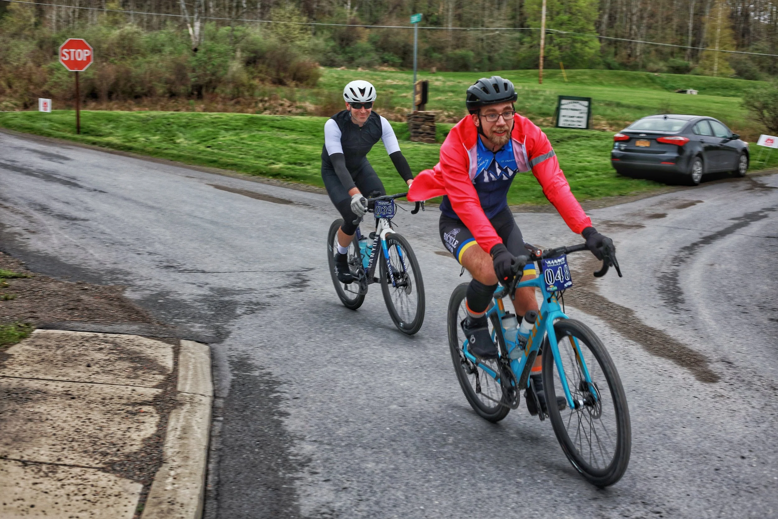 Two cyclists riding on a wet road near a stop sign and grassy area with trees in the background.
