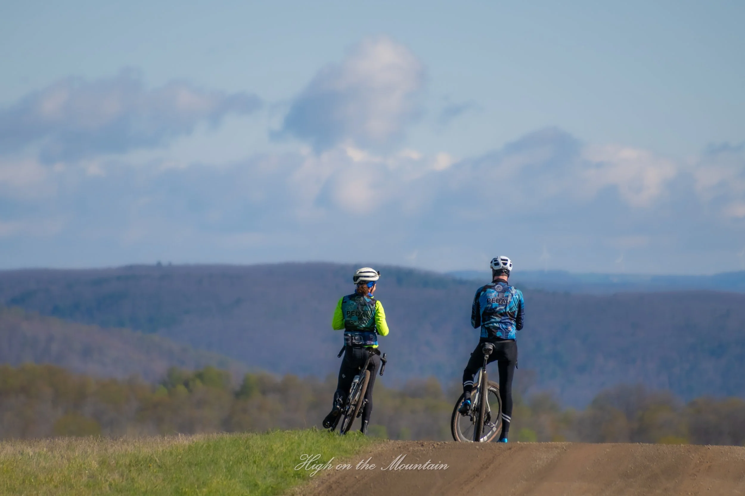 Two cyclists standing on a mountain trail, overlooking a distant landscape with a cloudy sky.