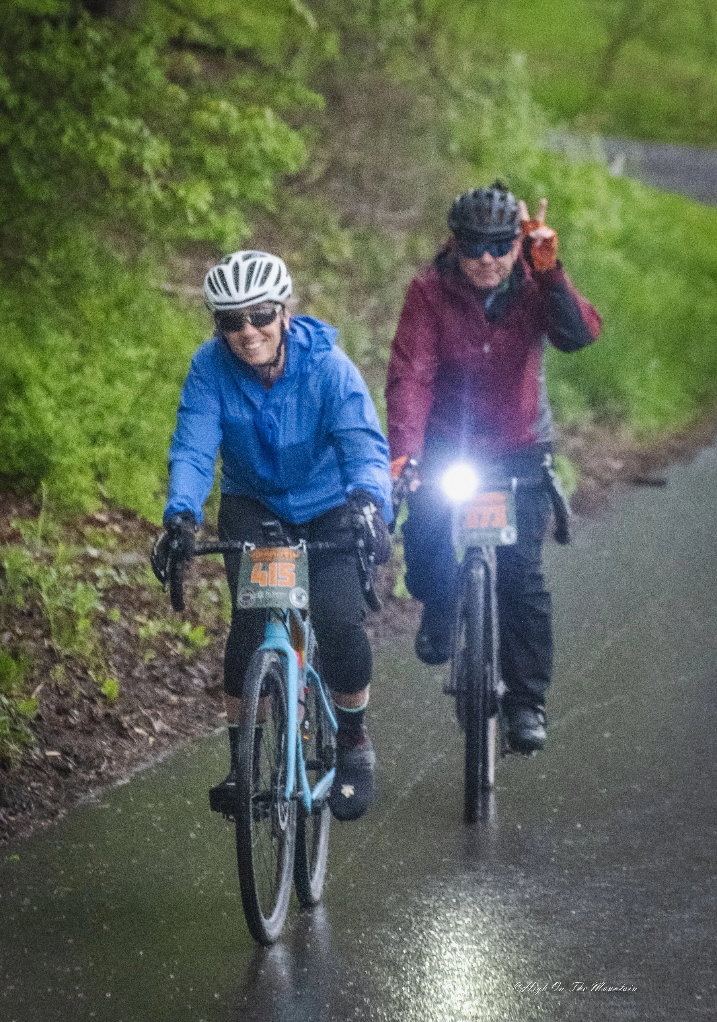 Two cyclists ride on a wet trail through a lush, green forest. Both are wearing helmets, sunglasses, and waterproof jackets; one in blue and the other in maroon. The cyclist in maroon is making a peace sign with their hand.