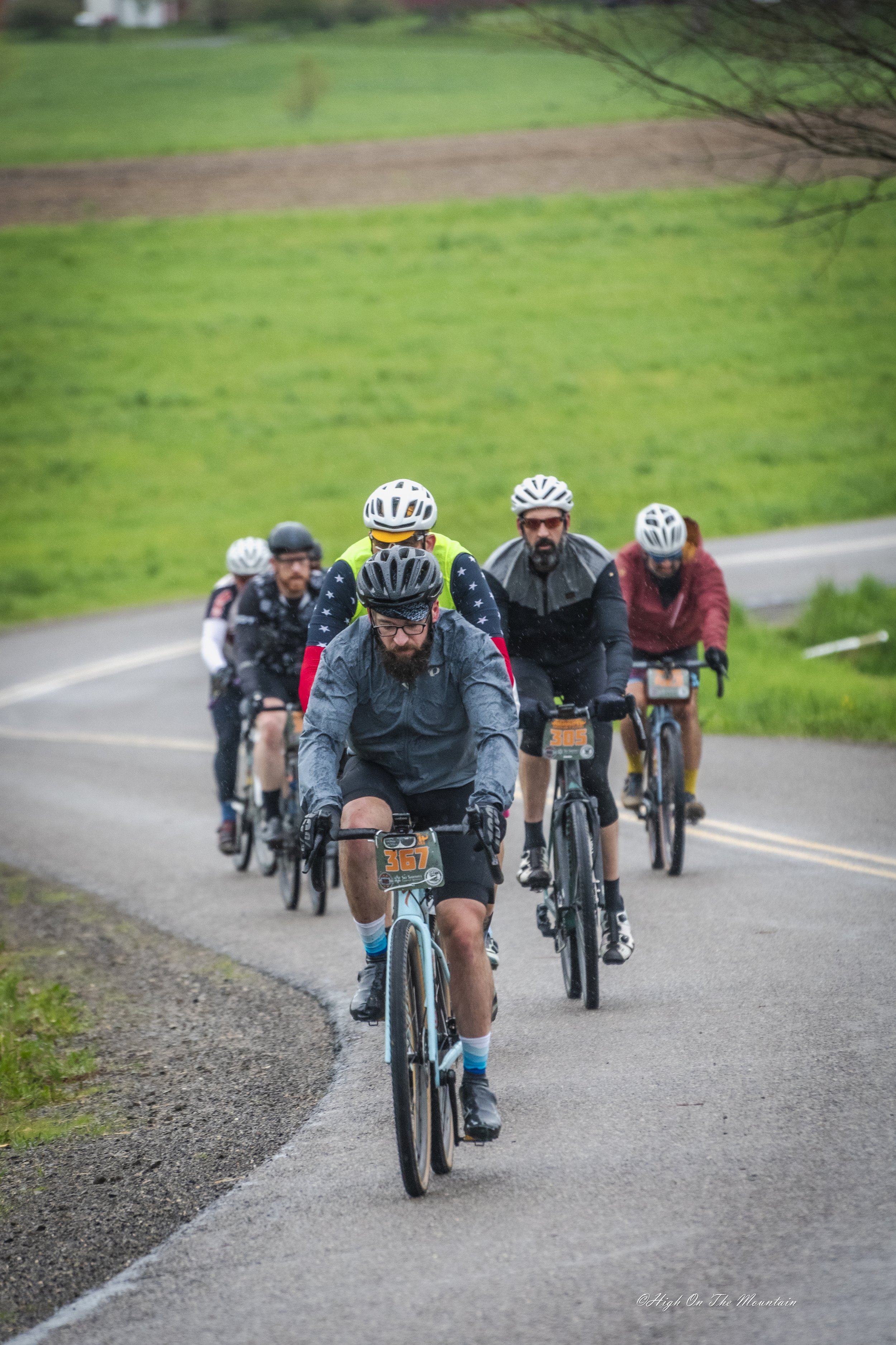 Group of cyclists riding on a paved trail through a green rural landscape with rolling hills and trees.