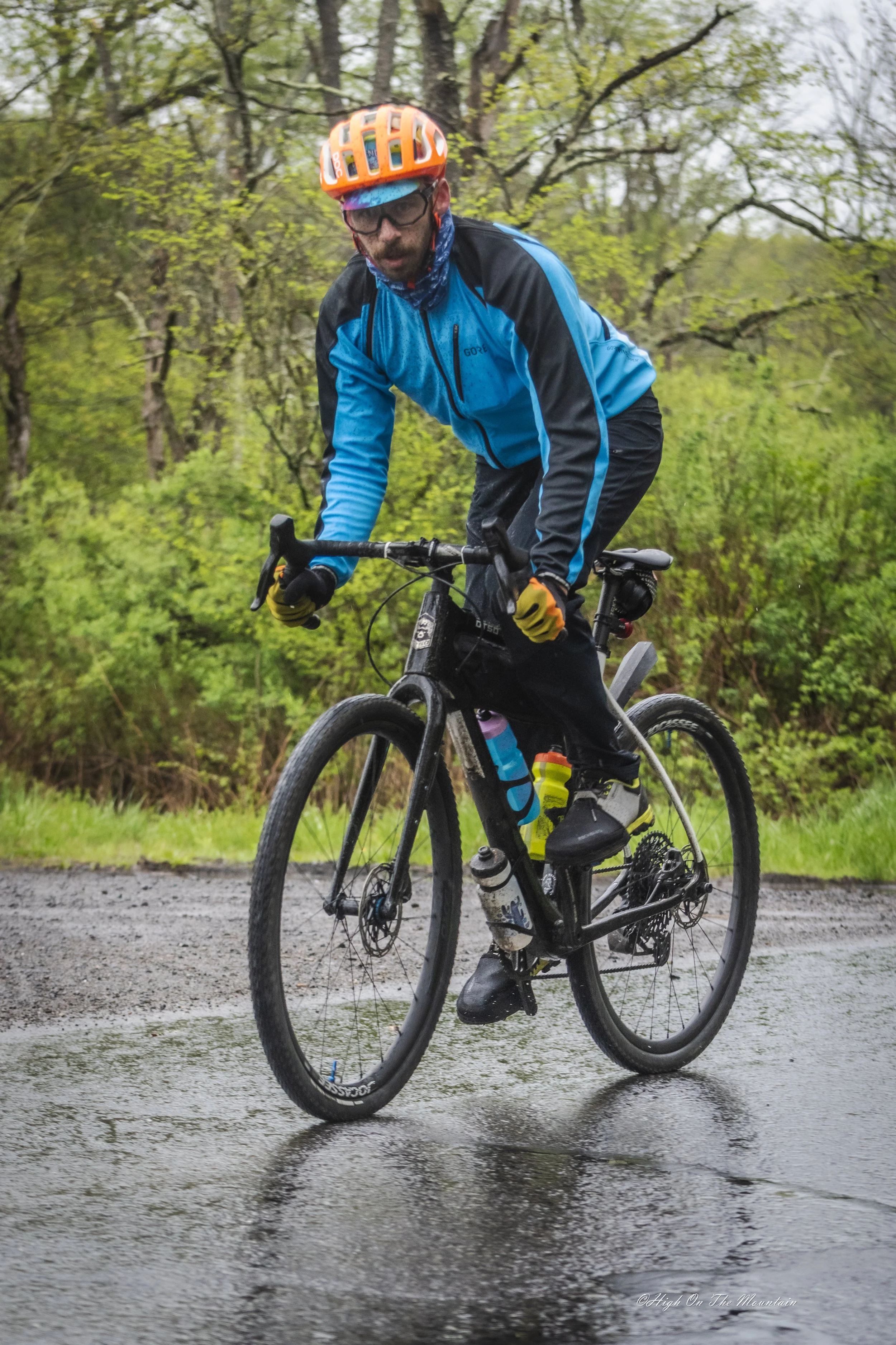 A man wearing an orange helmet, glasses, and a blue and black cycling jacket riding a mountain bike through a shallow water puddle on a dirt trail surrounded by green trees.