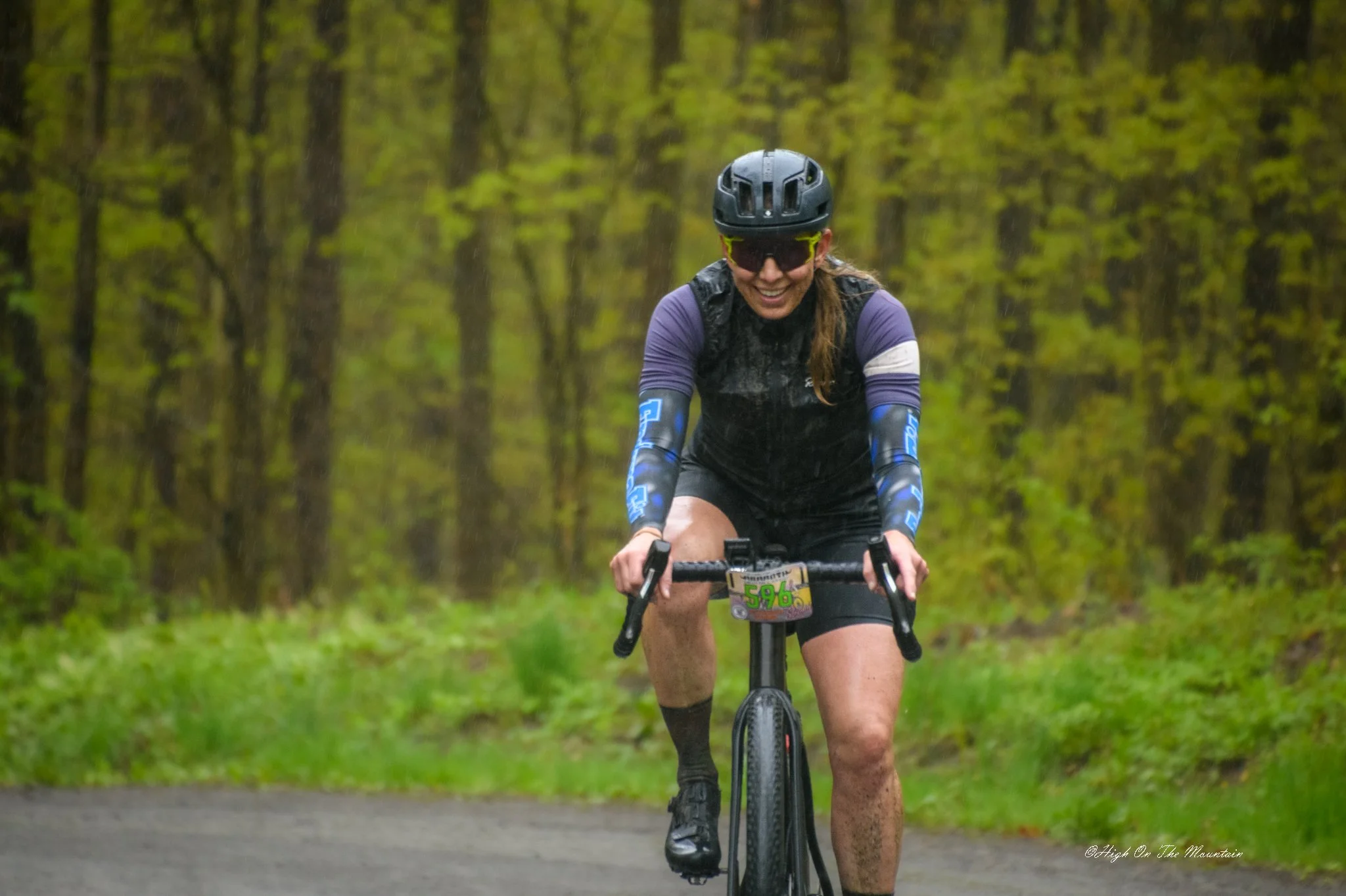 A woman riding a mountain bike on a forest trail in the rain, wearing a helmet, sunglasses, and athletic clothing.
