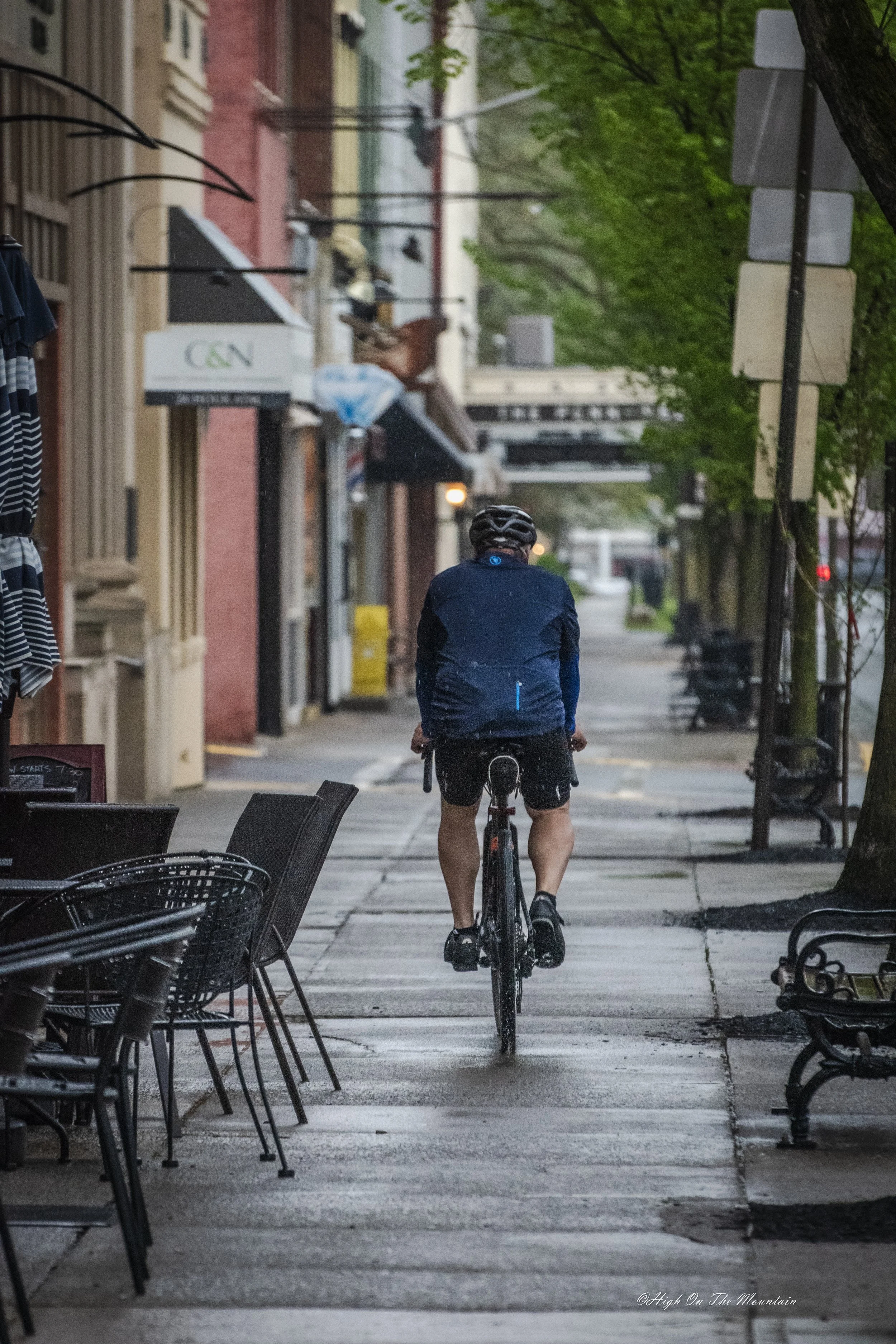 A man riding a bicycle on a city sidewalk during early morning or late evening, with outdoor seating and empty chairs on the side, trees with green foliage, and storefronts lining the street.