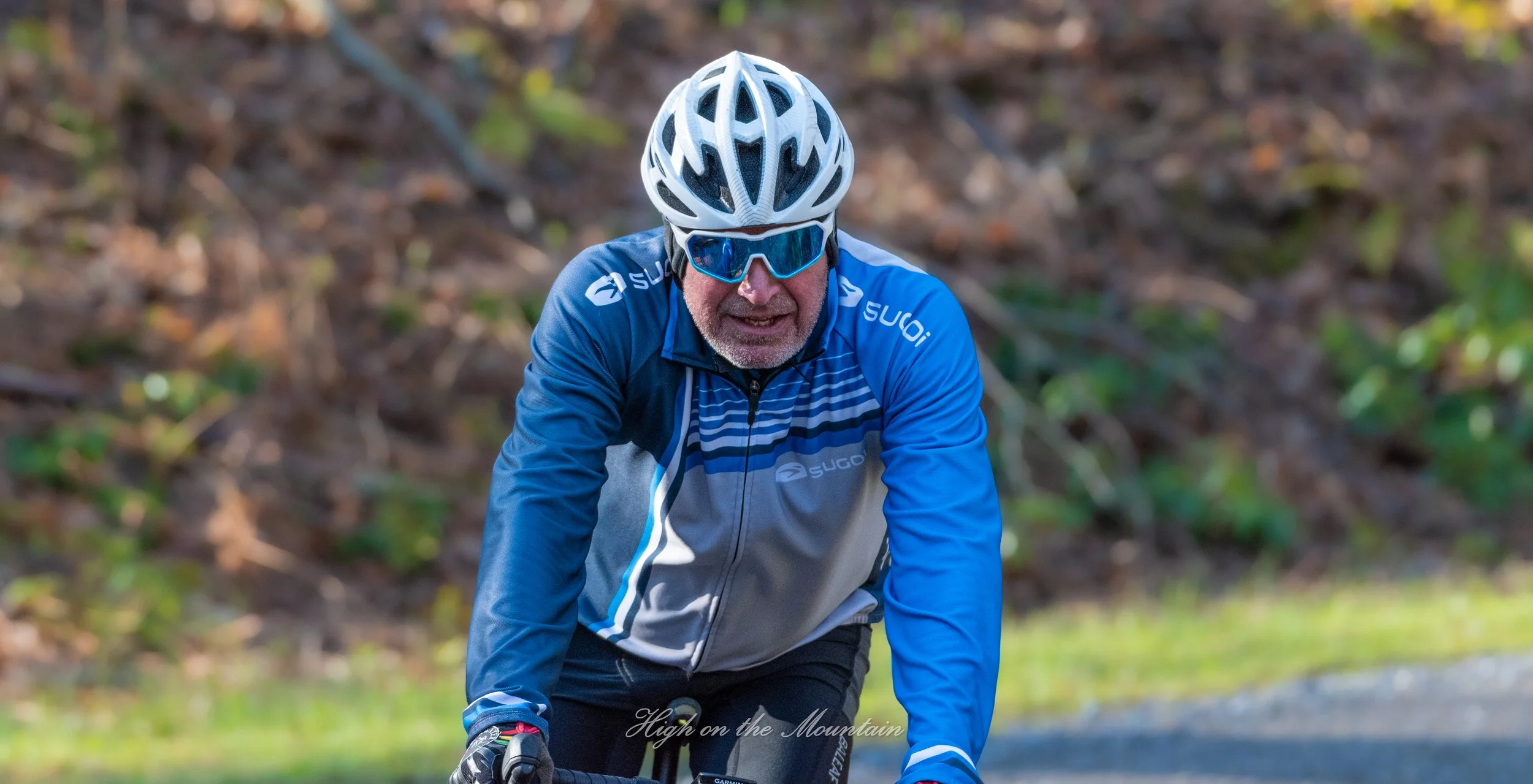 A man wearing a white bicycle helmet, blue sunglasses, and a blue and gray cycling jacket riding a bike outdoors on a trail surrounded by green foliage.