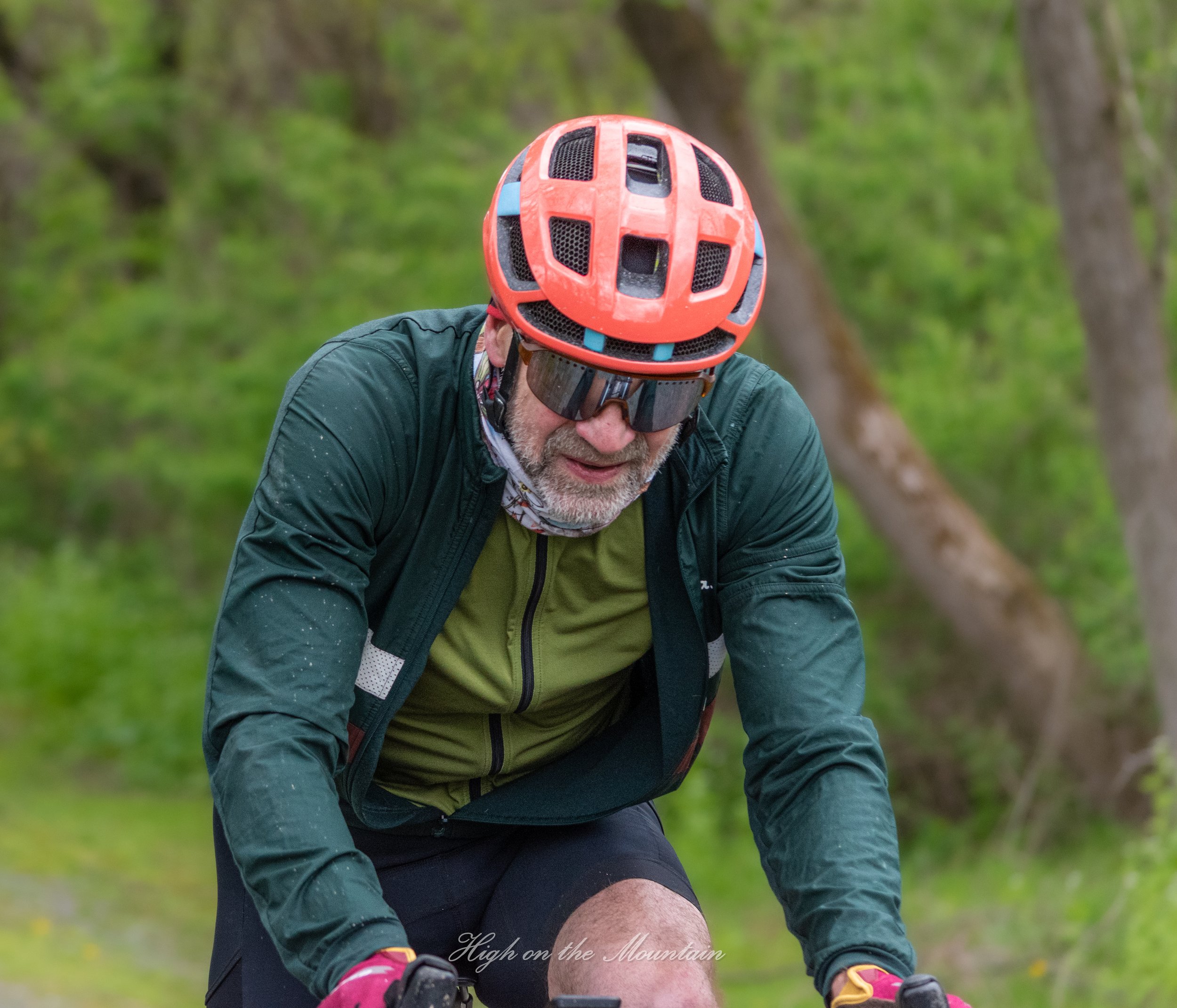 A man wearing a red helmet with black vents, sports sunglasses, a green jacket, and a patterned neck gaiter, riding a bike through a green forested trail.