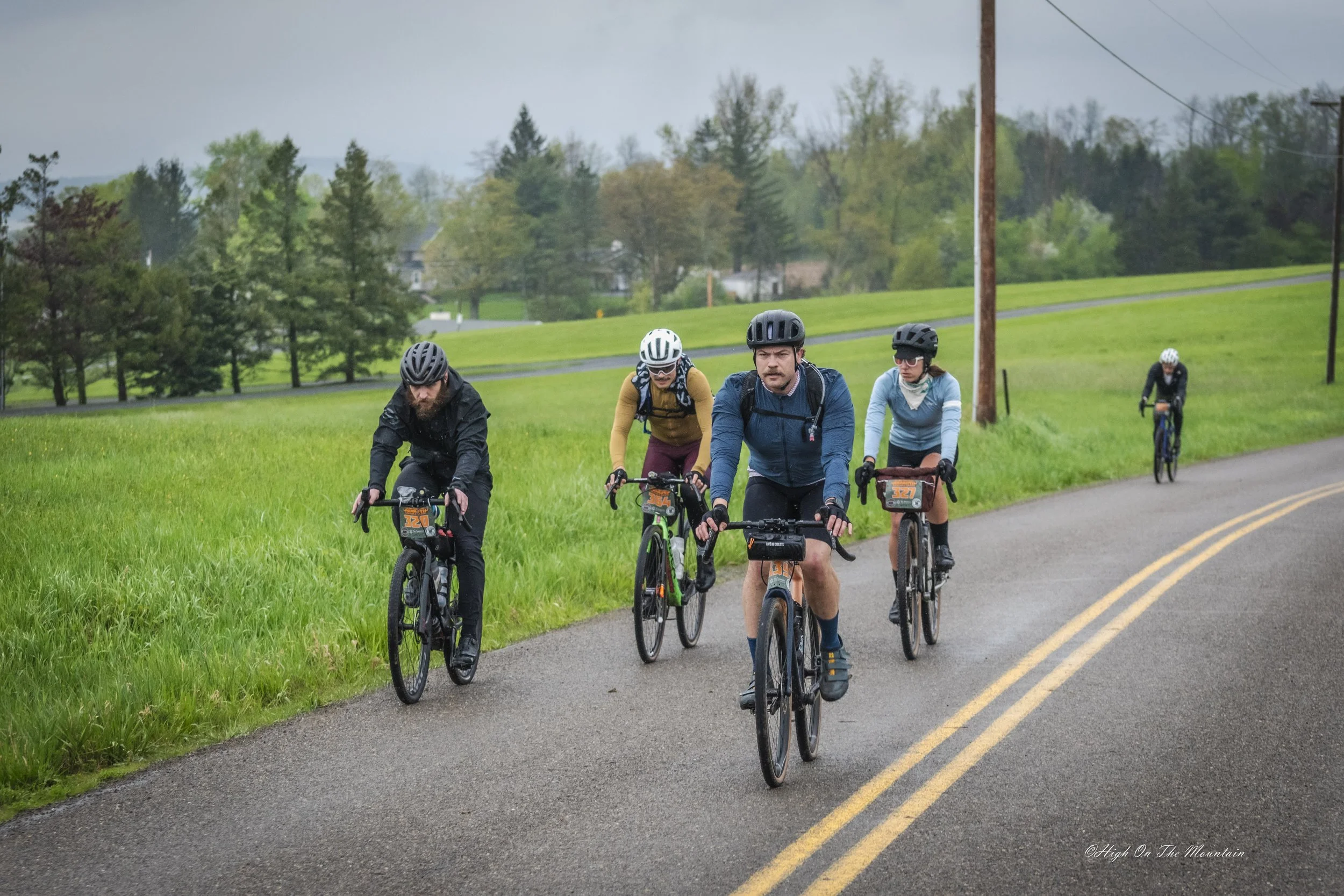Group of five cyclists riding on a paved road through a lush green landscape with trees and houses in the background, under an overcast sky.
