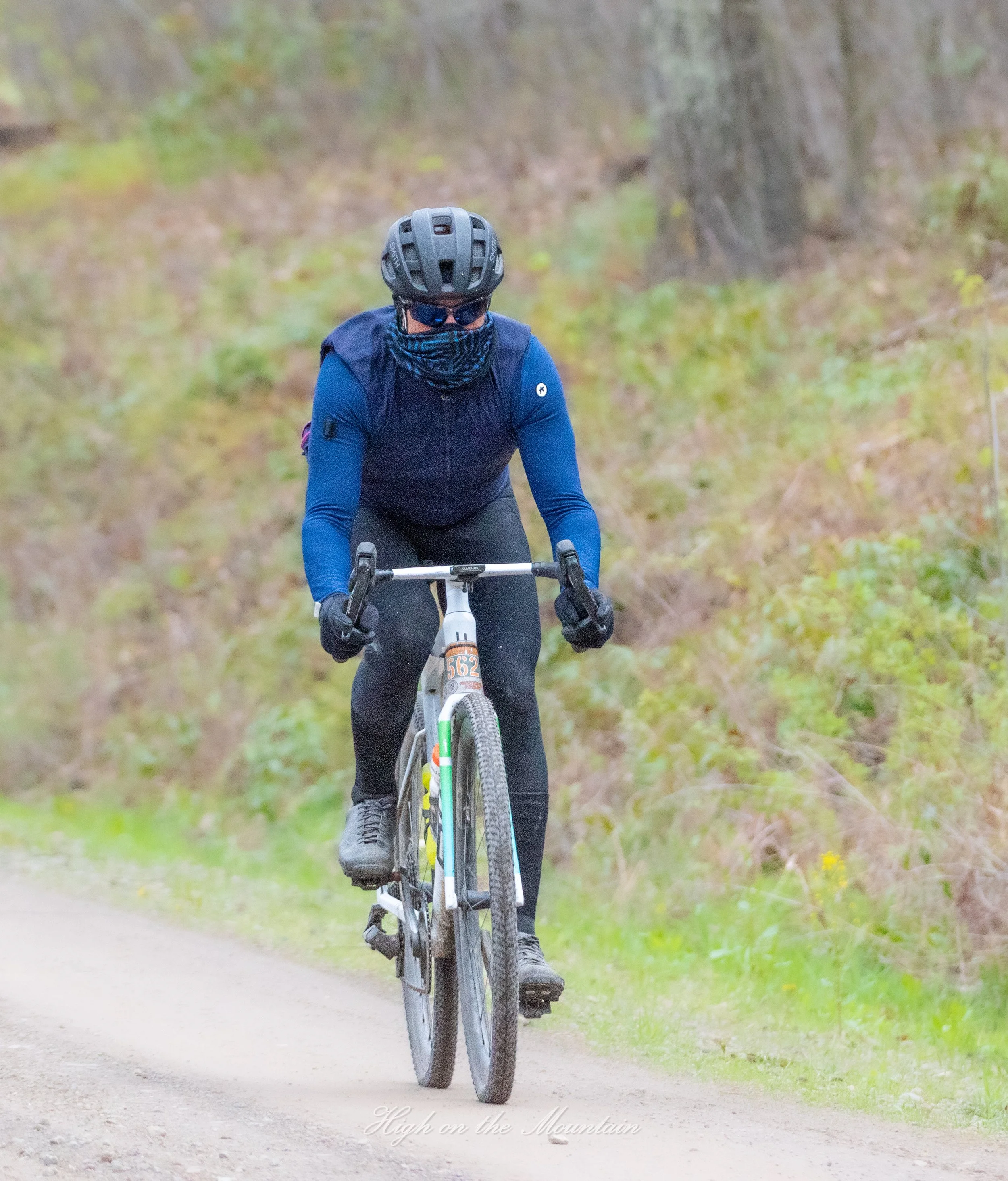 A person cycling on a dirt trail through a wooded area, wearing a navy blue long-sleeve shirt, black pants, a black helmet, sunglasses, and a face covering.