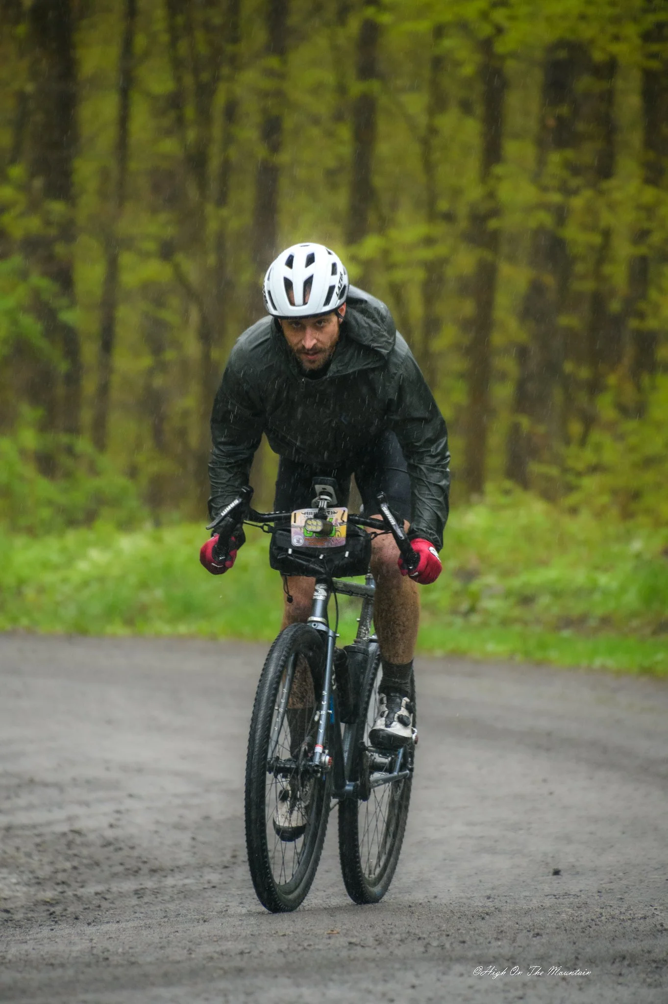 A man wearing a white helmet rides a mountain bike on a dirt trail in a wooded area during rain, wearing a black rain jacket and red gloves.