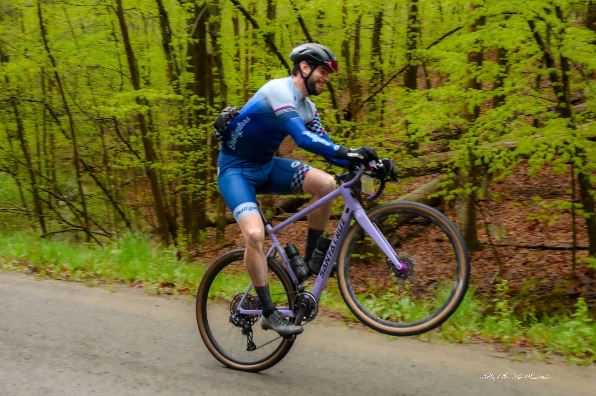 A man biking on a mountain trail through a lush green forest, wearing a helmet and sports gear, smiling as he rides a purple bicycle.