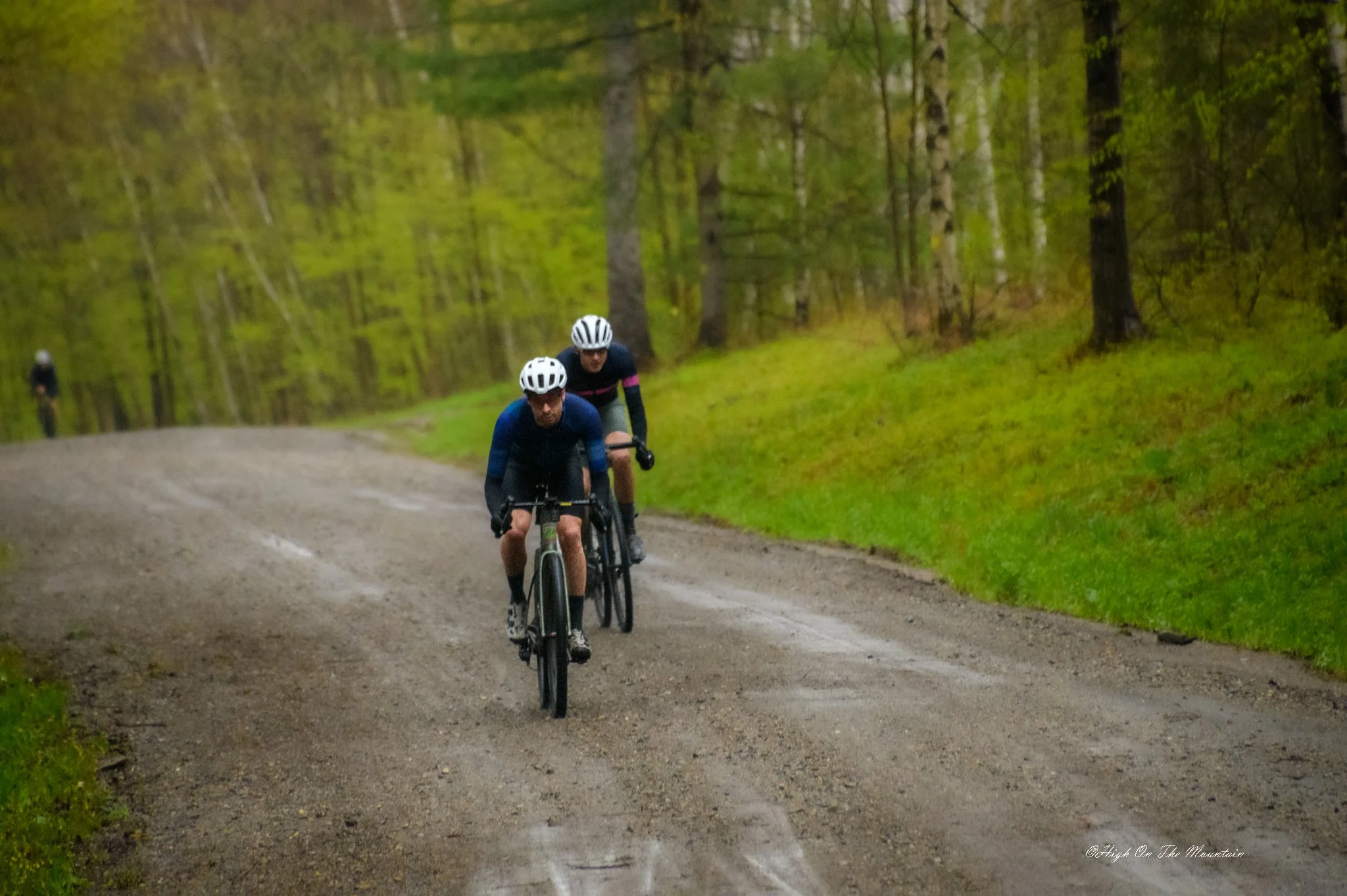 Two men riding bicycles on a dirt trail through a forest with green trees, with a third cyclist visible in the background.