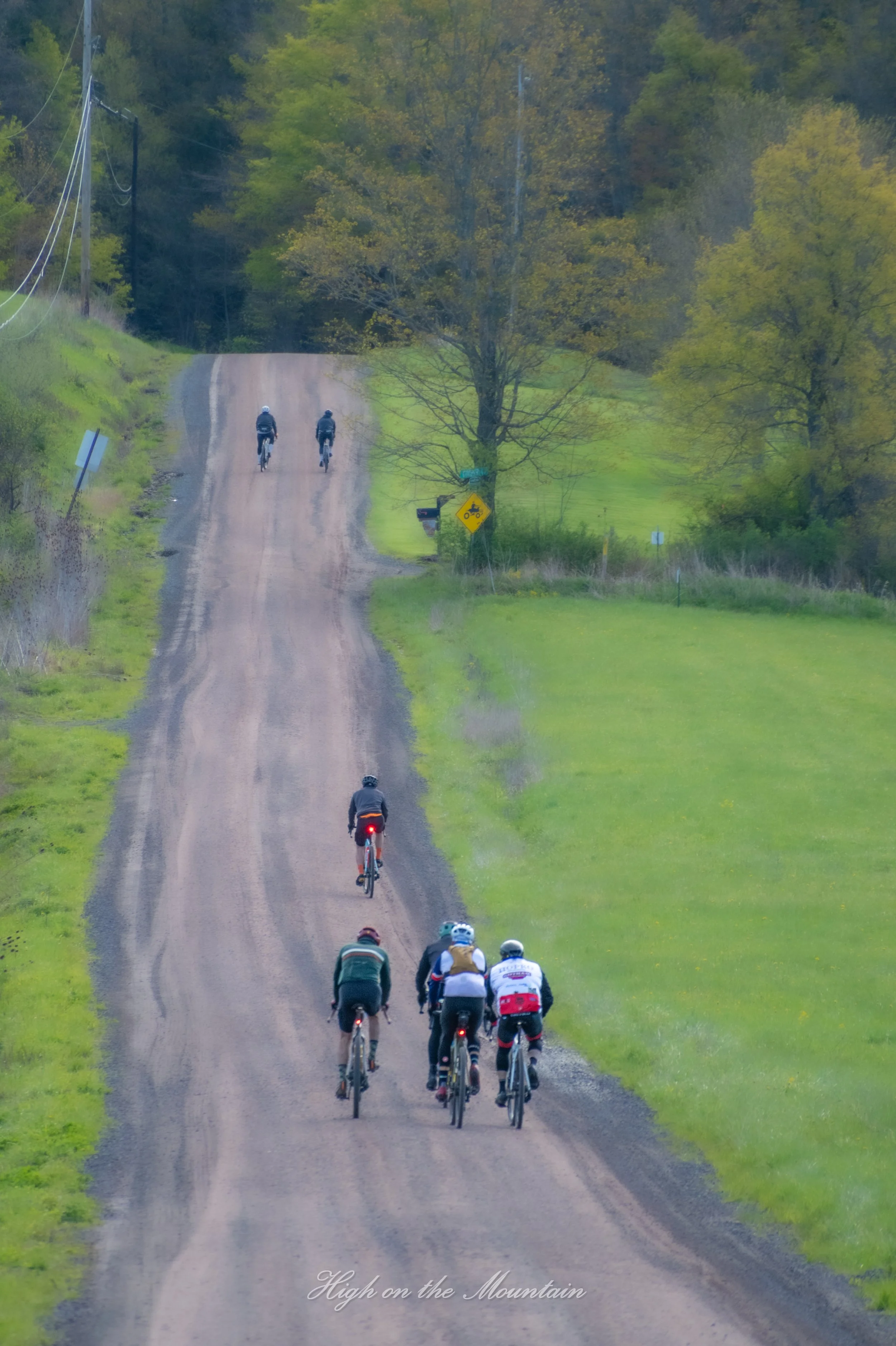 A group of seven cyclists riding on a rural dirt road surrounded by green trees and grass, with utility poles and a road sign visible along the roadside. Text at the bottom reads 'High on the Mountain'.