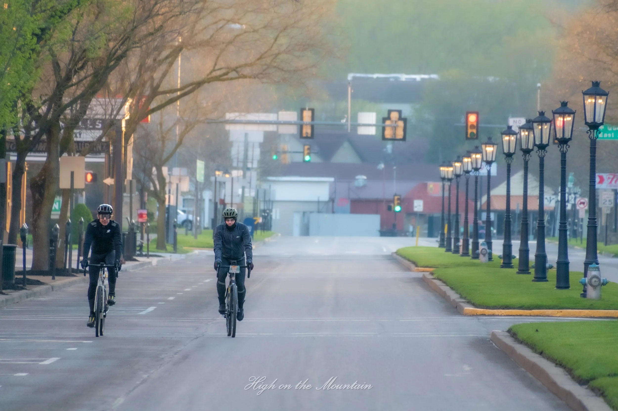 Two men riding bicycles on a street lined with decorative street lamps, trees, and buildings, with traffic lights in the background, early morning or late afternoon lighting, and fog in the distance.