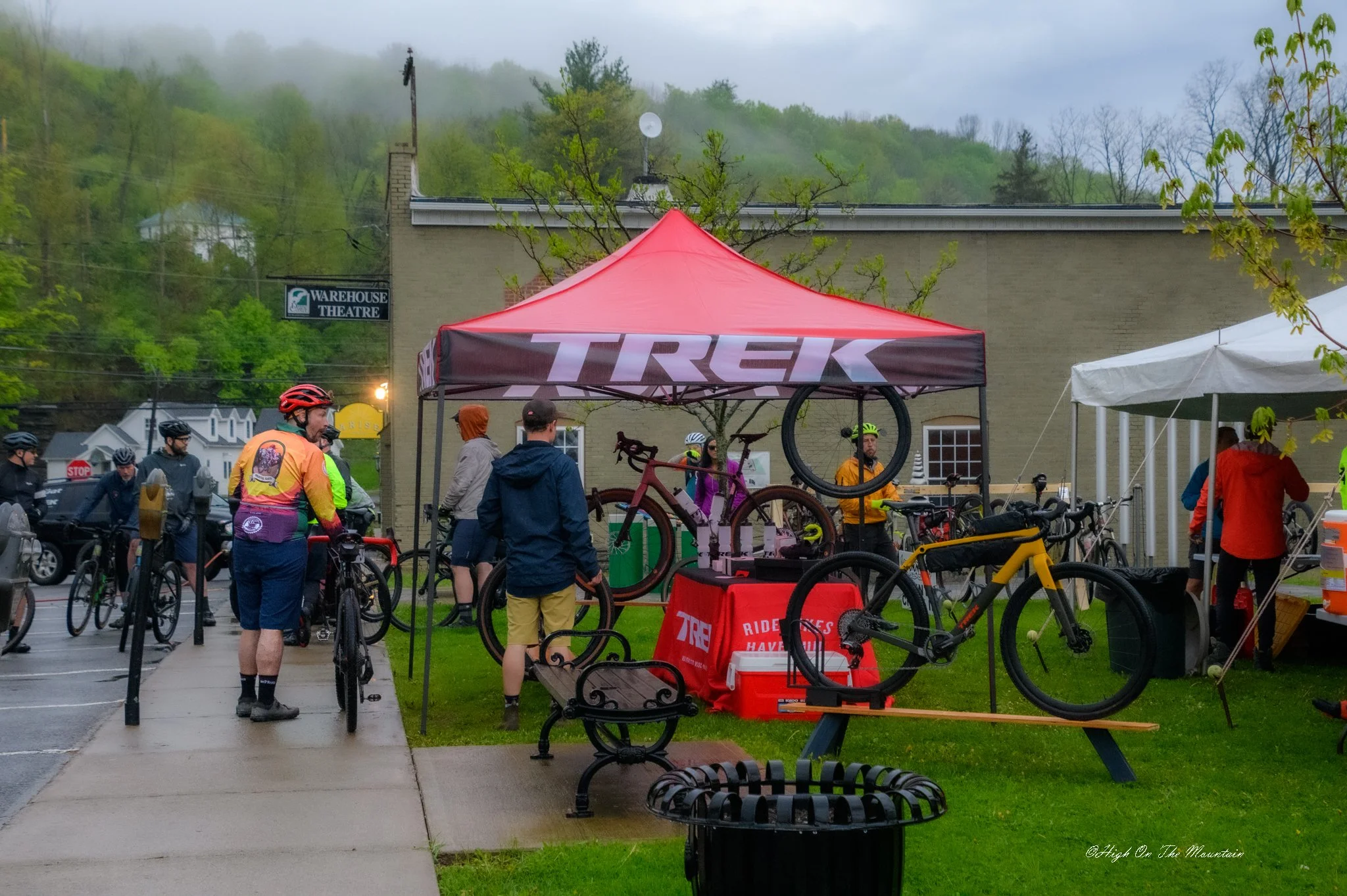 Outdoor event where people gather around a red tent with the Trek logo, displaying bicycles and cycling gear, with a backdrop of a building and a foggy, green, hilly landscape.