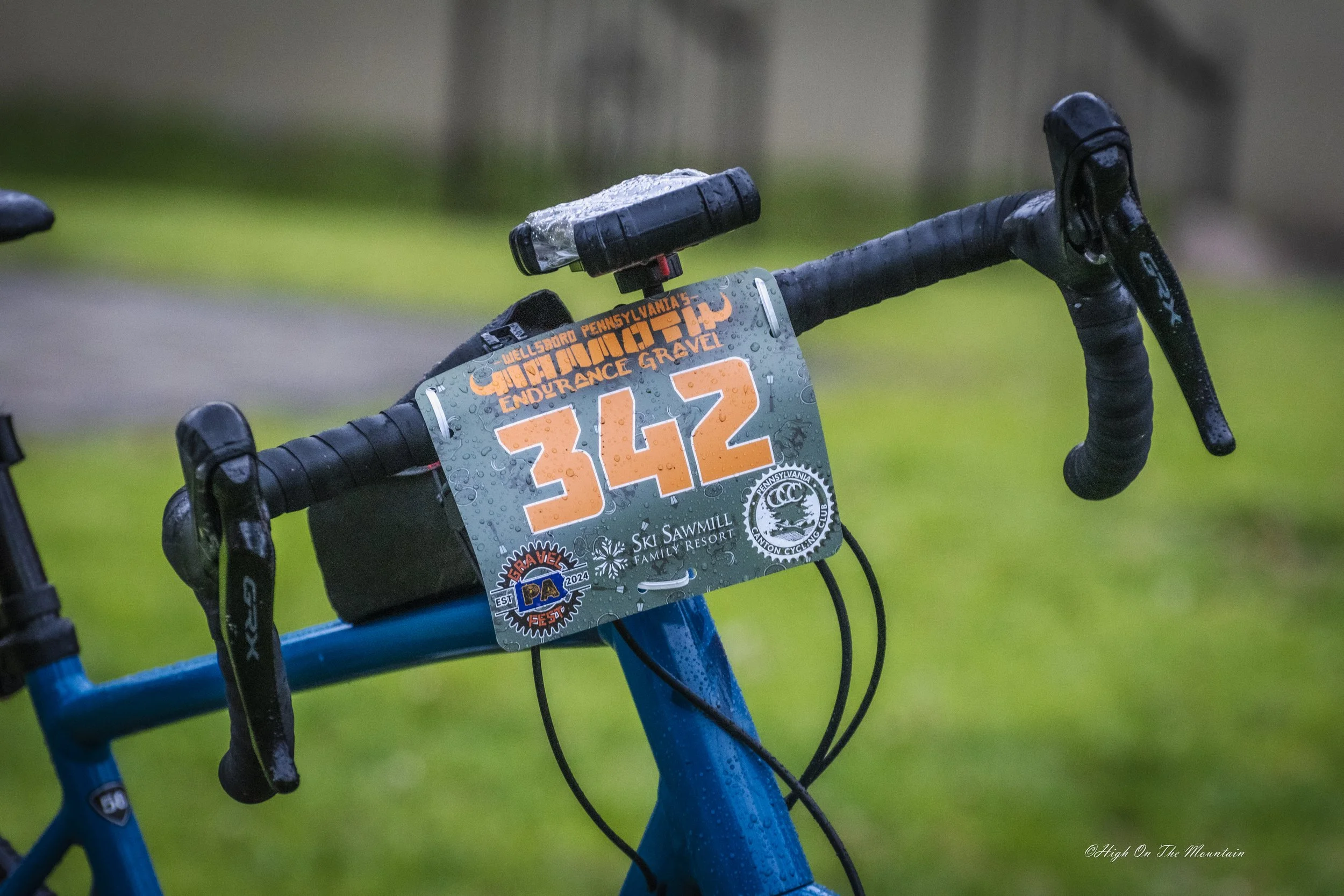 Close-up of a blue bicycle handlebar with a race number 342 attached, wet from rain, set against a grassy background.