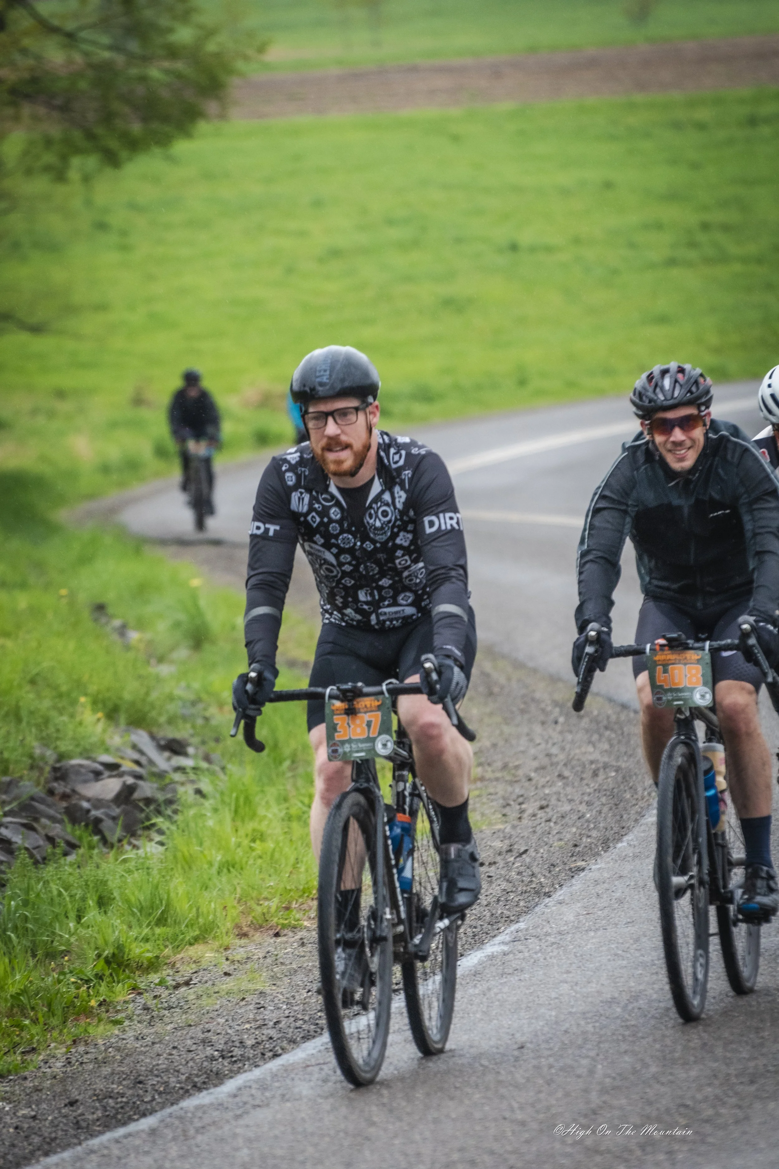 Two male cyclists riding on a paved trail in a green outdoor setting with grassy fields and trees, wearing helmets and cycling gear.