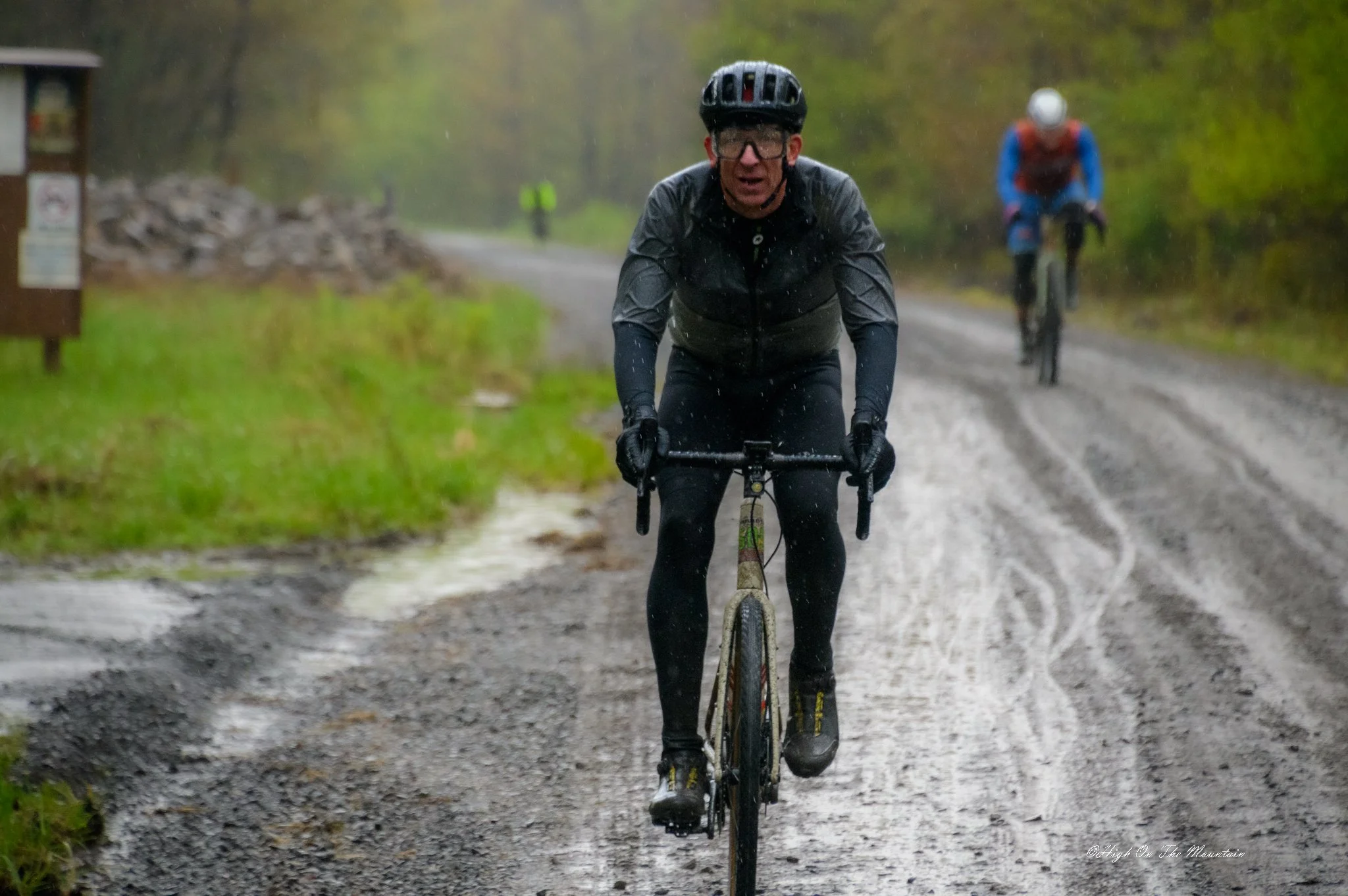 A man riding a bicycle in the rain on a muddy trail with another cyclist in the background, wearing rain gear and a helmet.