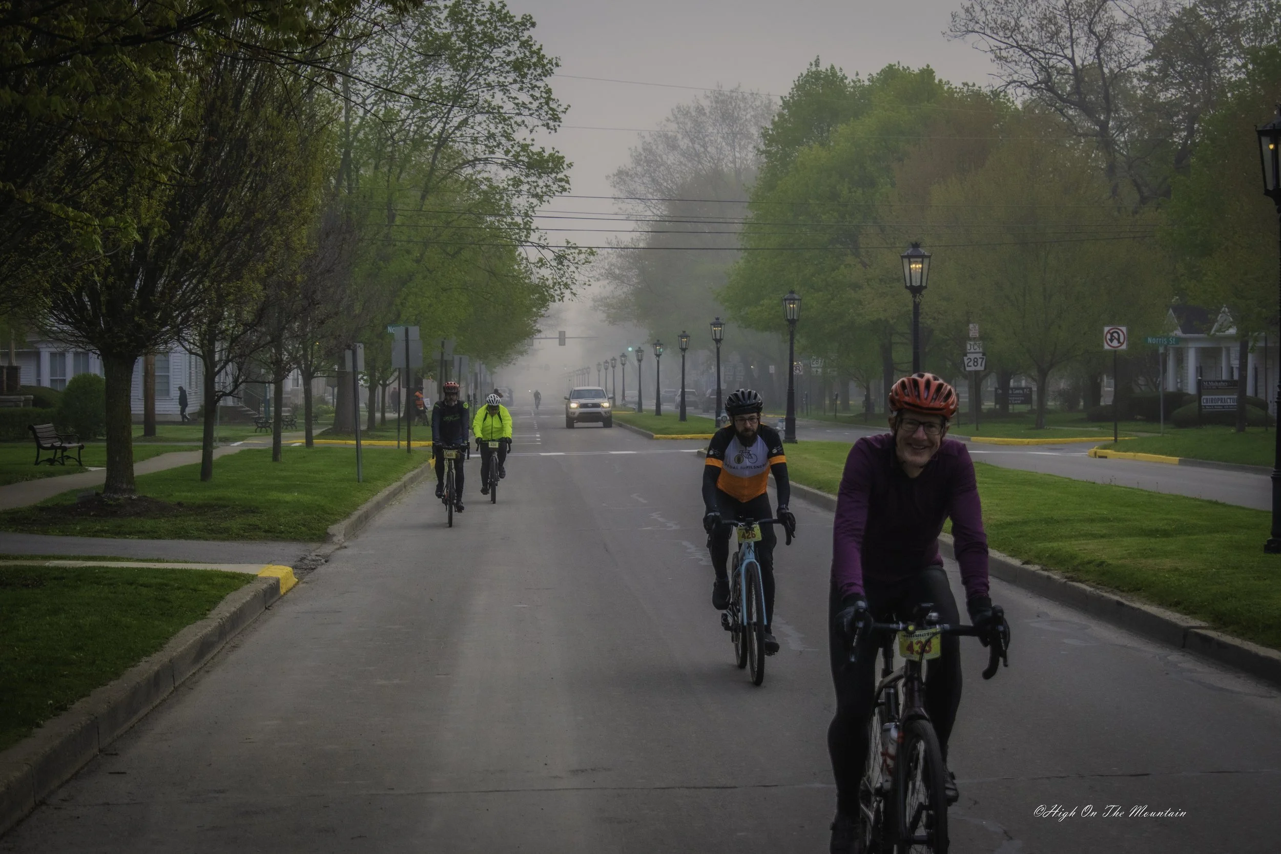 People riding bicycles on a foggy street lined with trees and lamp posts in a residential neighborhood.