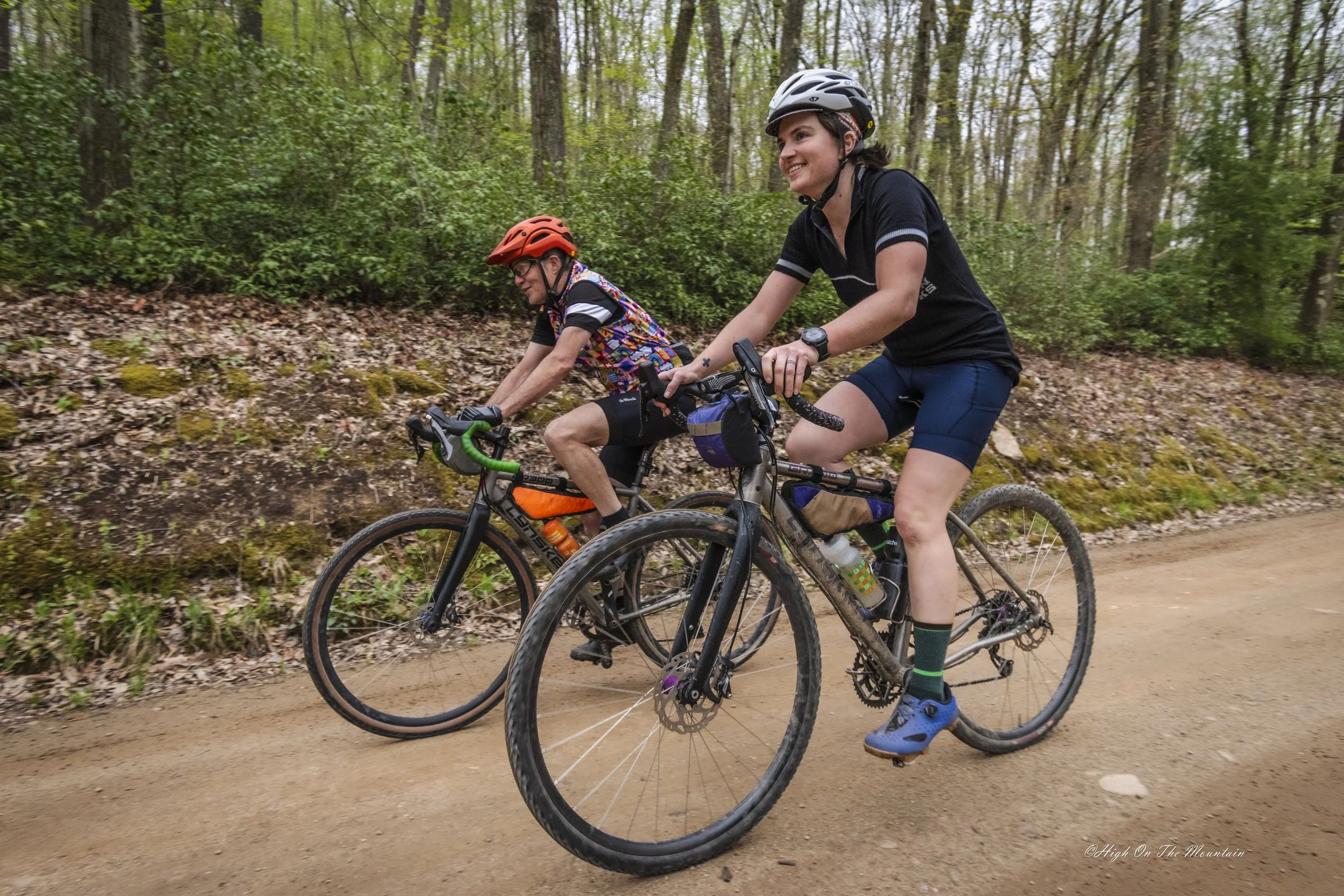 Two people riding mountain bikes on a dirt trail through a forest. Both are wearing helmets, and the person in the foreground is smiling while looking ahead.