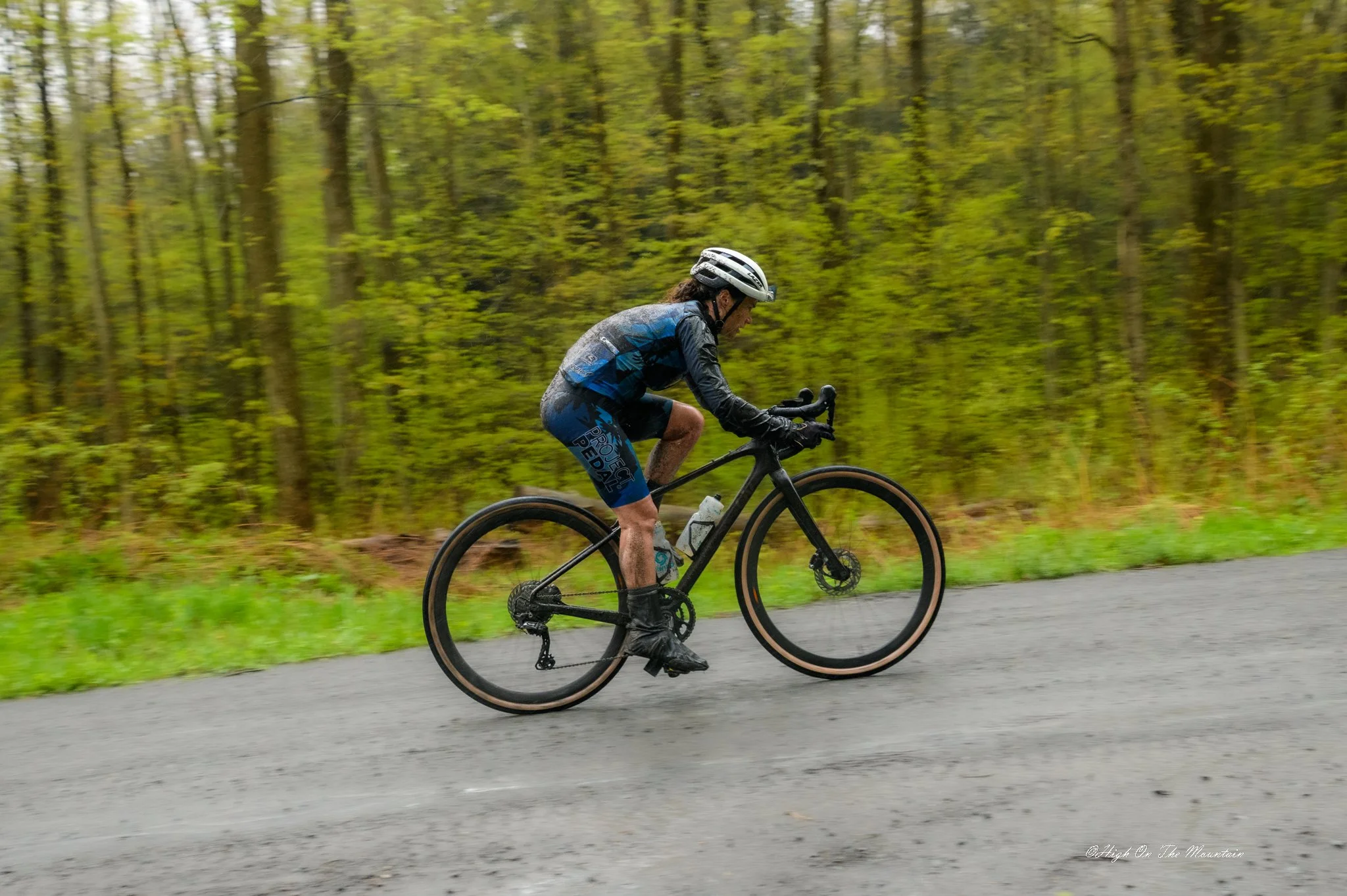 A man wearing a white helmet and cycling gear is riding a black mountain bike on a paved trail through a green forest.