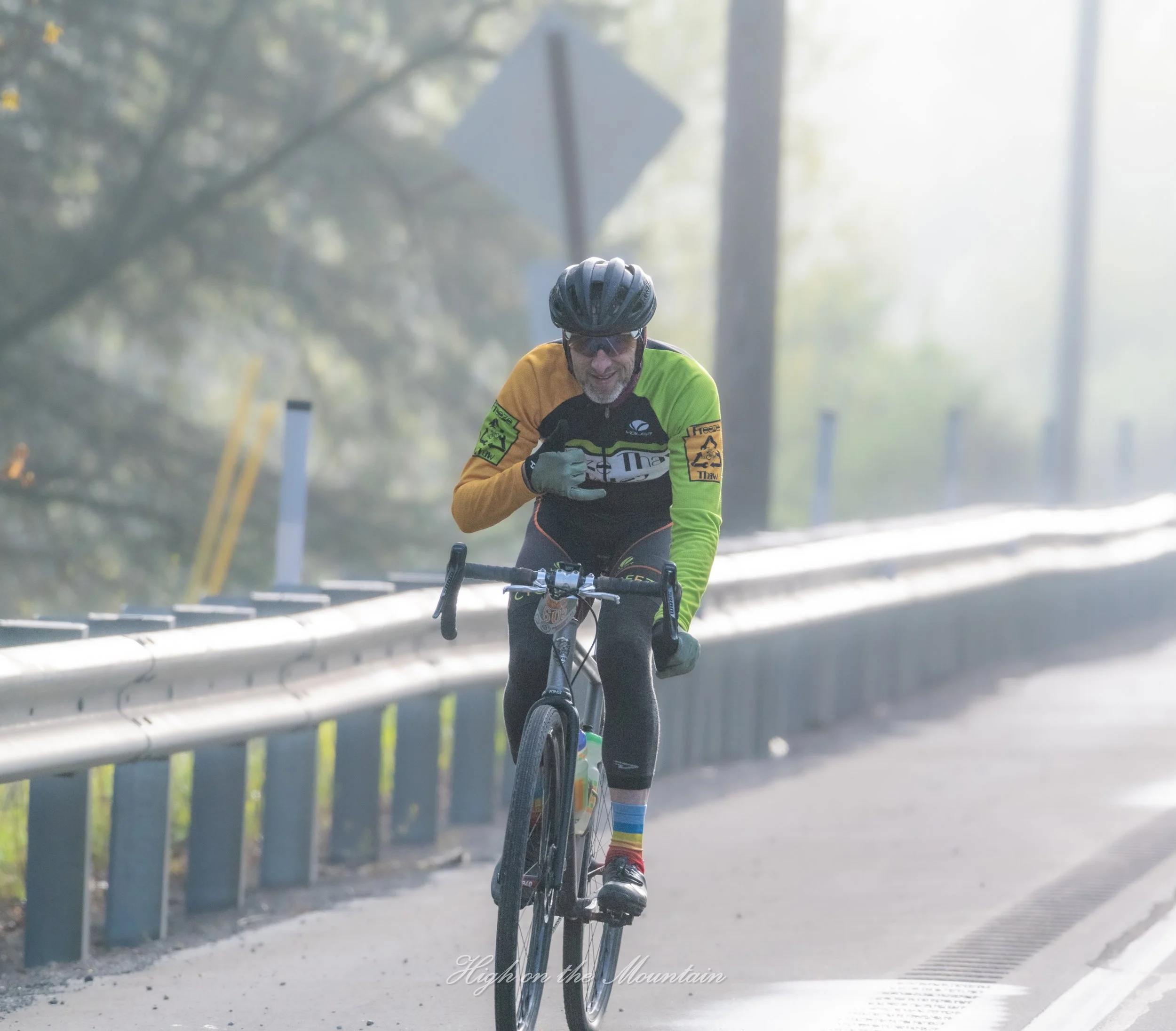 A cyclist wearing a black helmet, sunglasses, and a yellow and black cycling jersey riding on a road bordered by a metal guardrail and trees with sunlight filtering through.