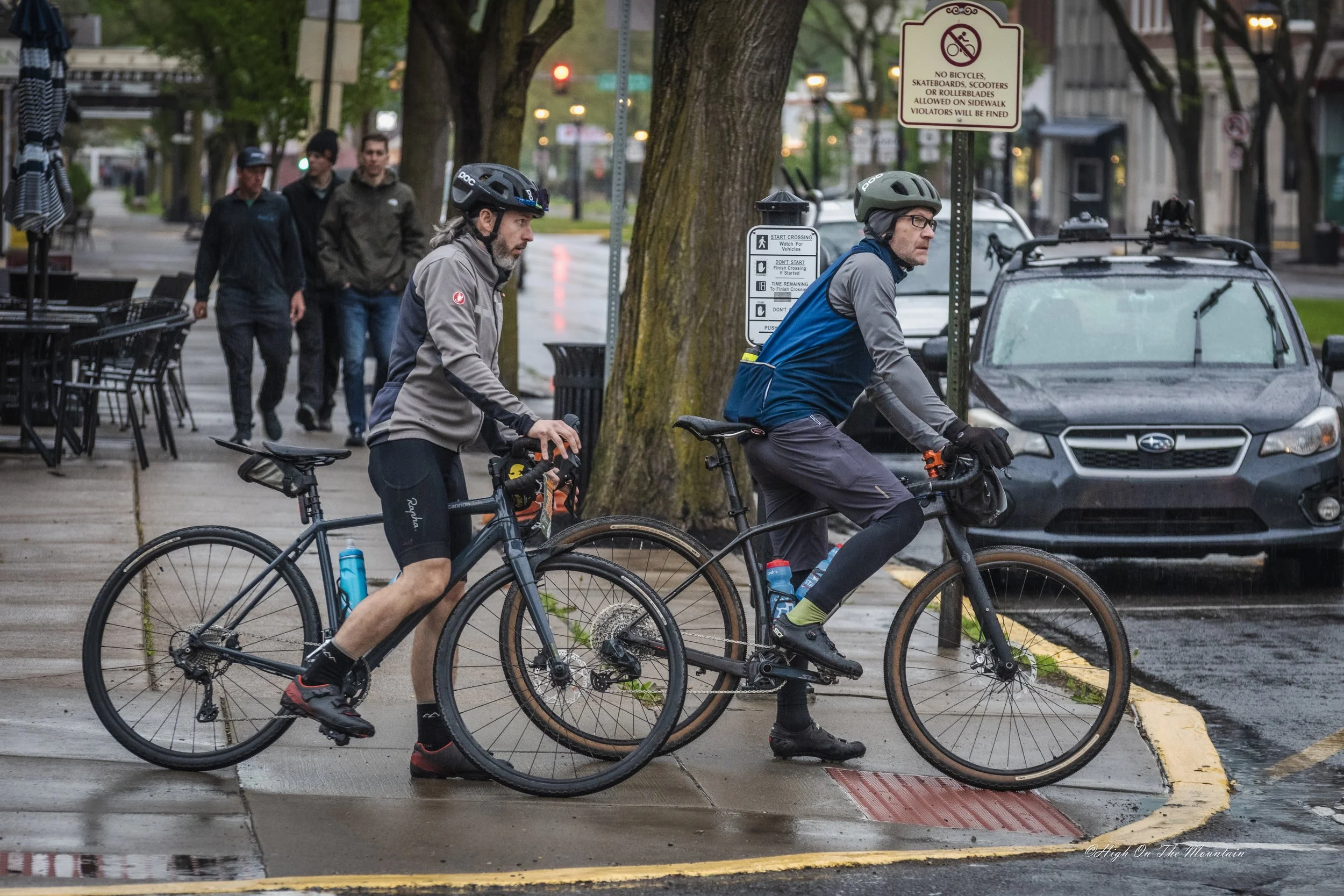 Two men wearing helmets riding bicycles on a city sidewalk, with pedestrians walking in the background and a sign about bicyclist rules nearby.