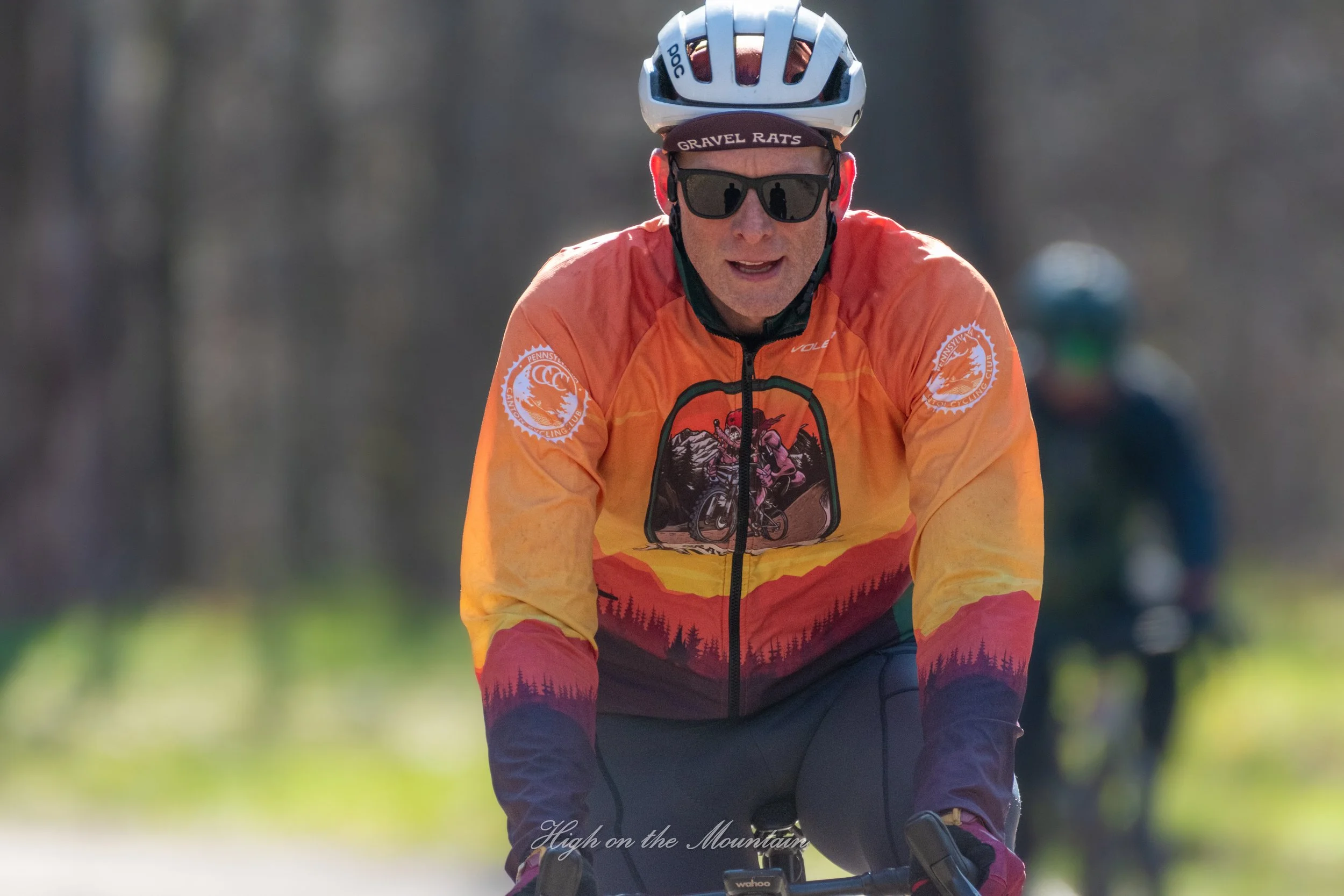A person riding a bicycle outdoors wearing sunglasses, a helmet, and a colorful jacket in shades of orange, yellow, and red with mountain and forest designs. The background is blurred, showing trees and another cyclist in the distance.