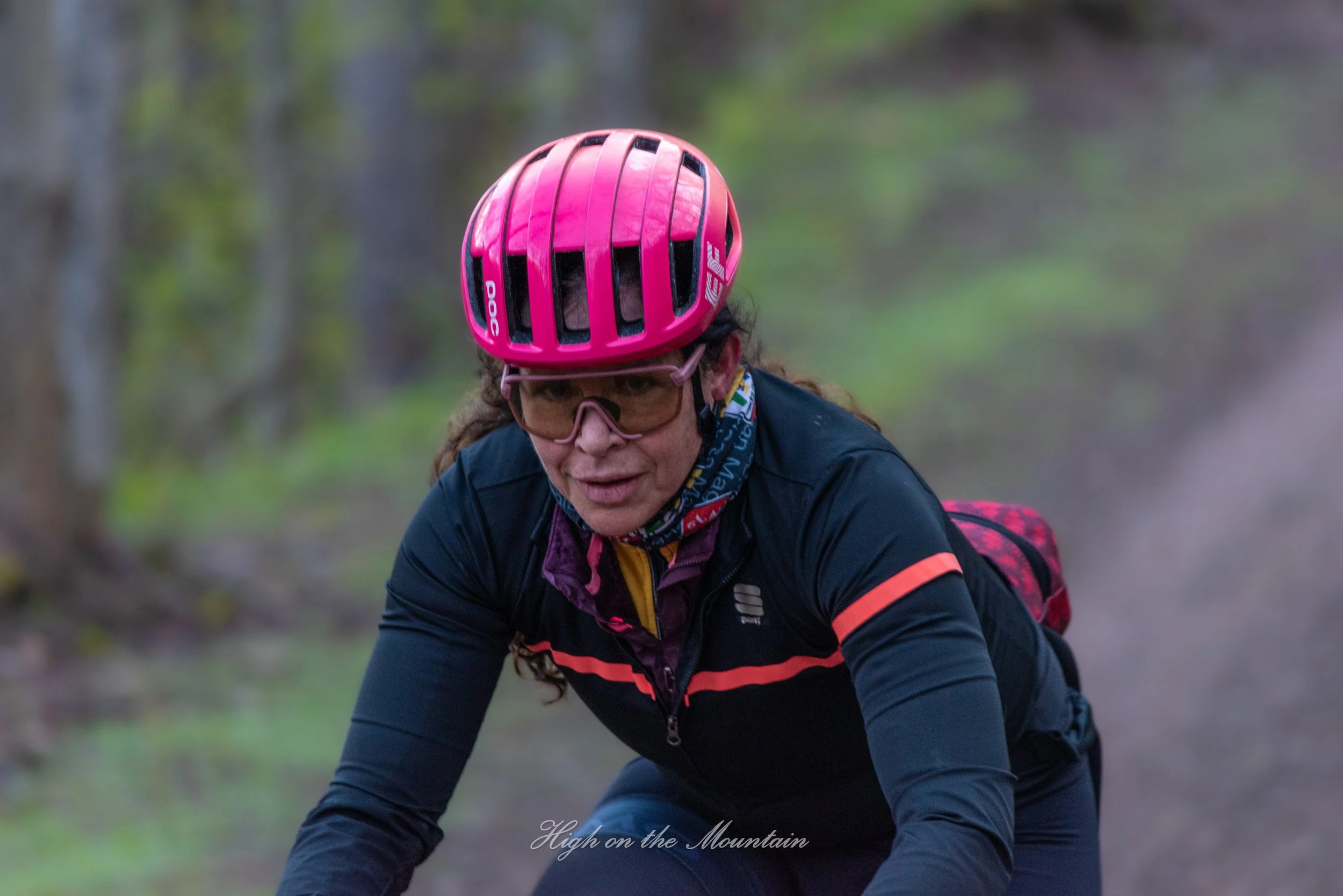 A woman in a pink bike helmet and black outdoor jacket riding a mountain bike on a forest trail.