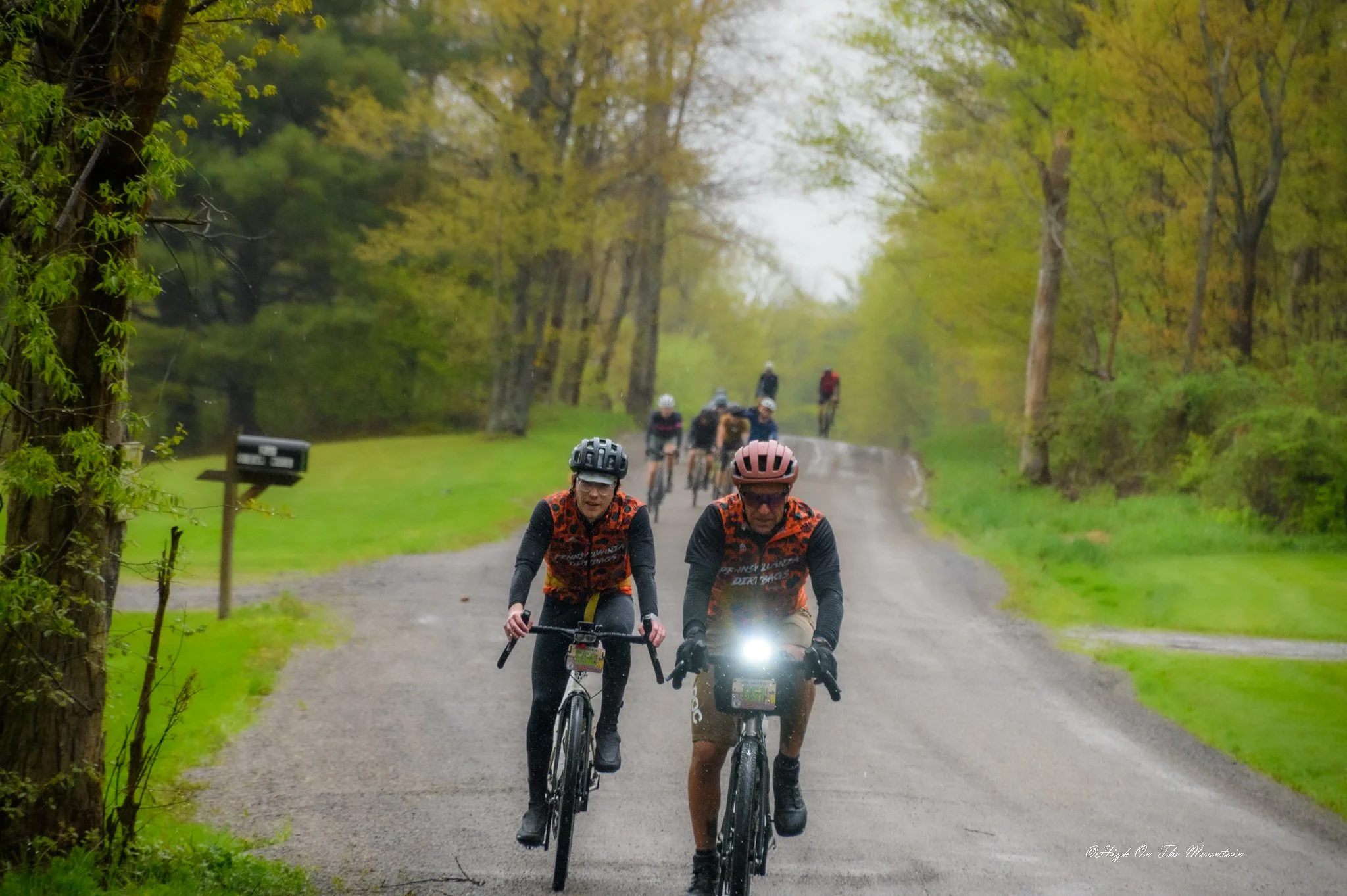 Group of cyclists riding on a forest trail on a rainy day, surrounded by green trees.