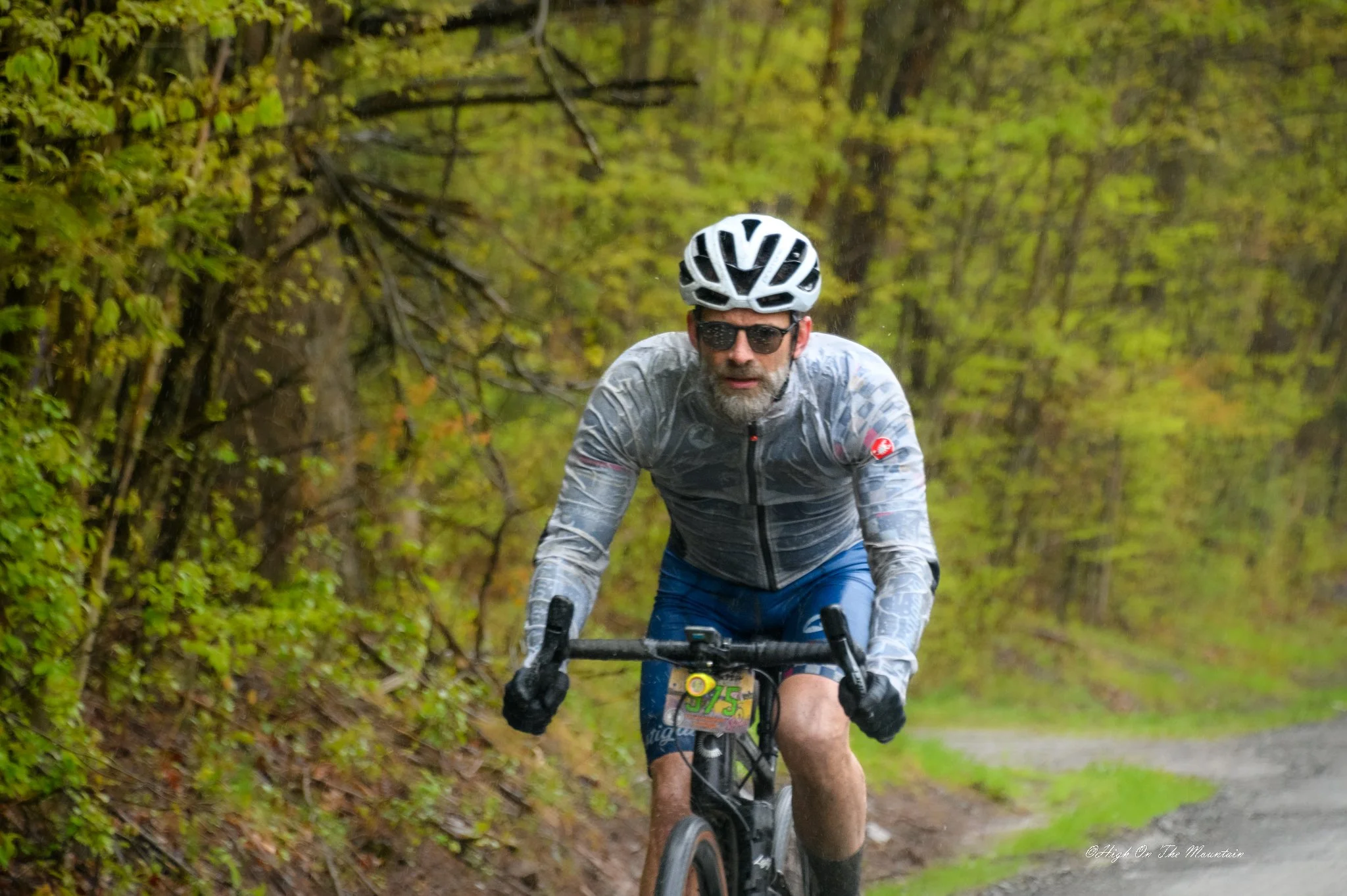 A man riding a mountain bike through a forest trail, wearing a helmet and sunglasses, surrounded by green foliage.