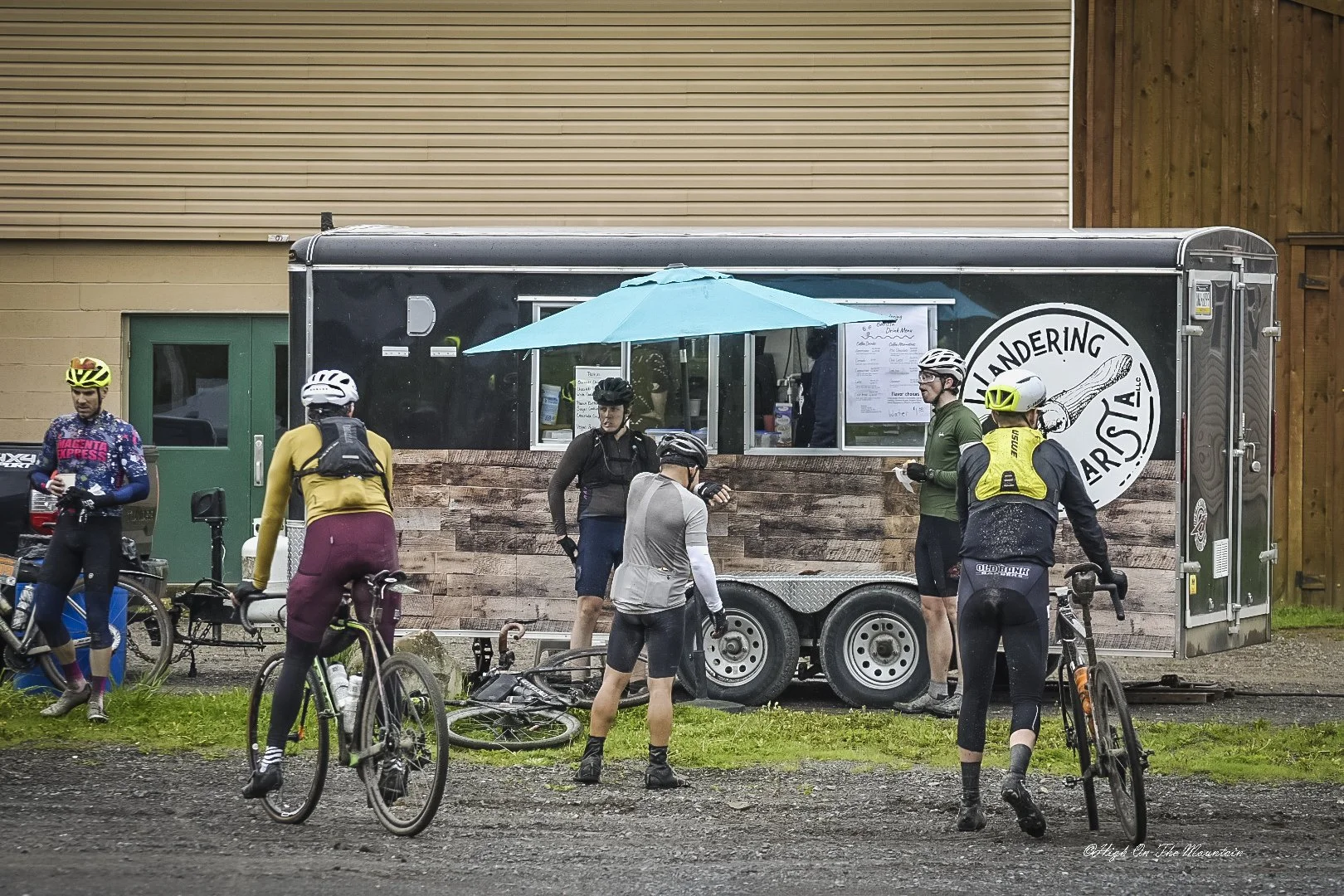Cyclists gather outside a black food truck with a large logo that says 'Wandering Arrow LLC' and an arrow pointing to the right. Some cyclists are standing with their bikes, chatting, while others are near a blue umbrella attached to the food truck. 