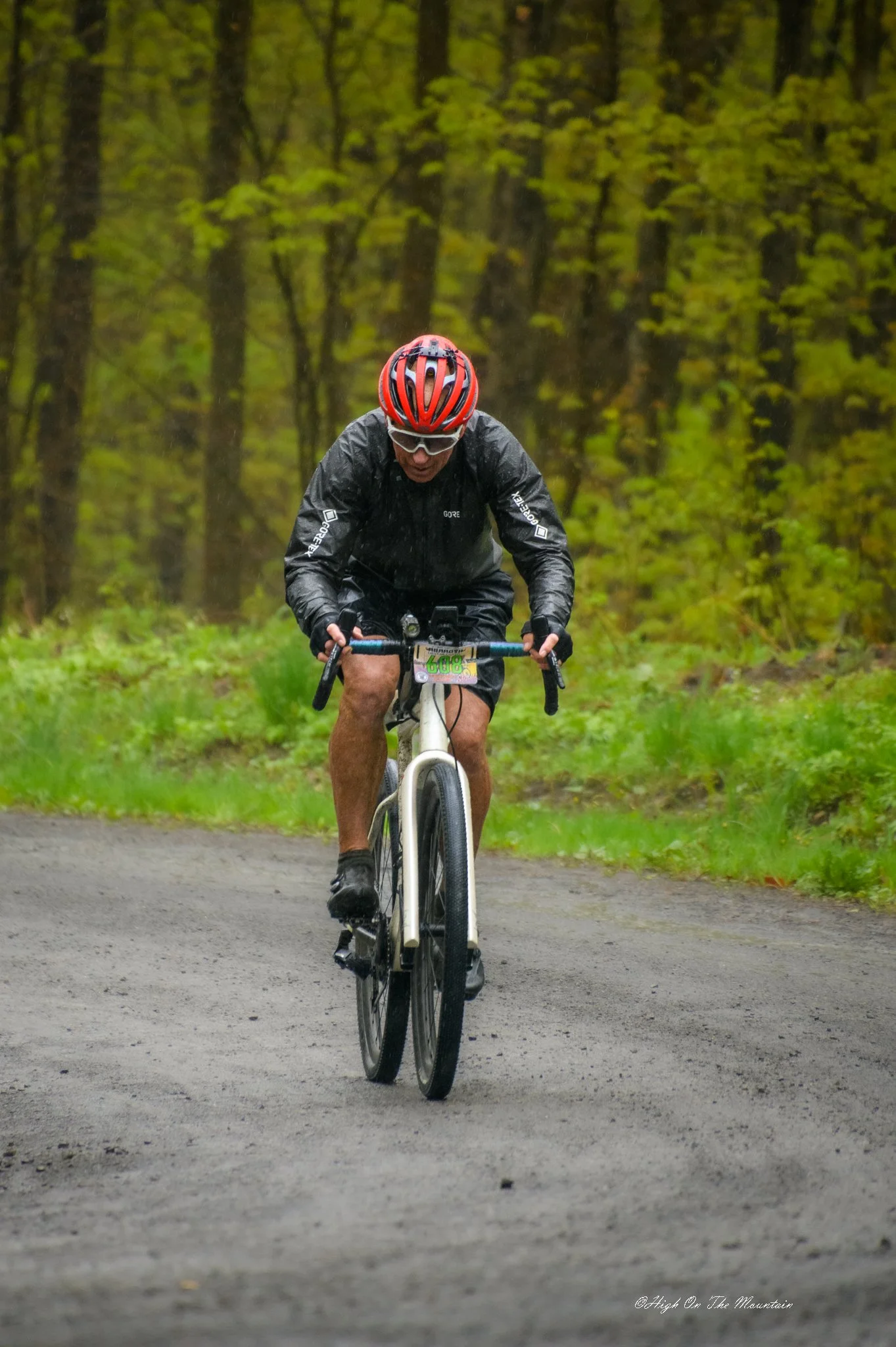 A person riding a mountain bike on a forest trail, wearing a red helmet, sunglasses, and a black rain jacket, with green trees in the background.