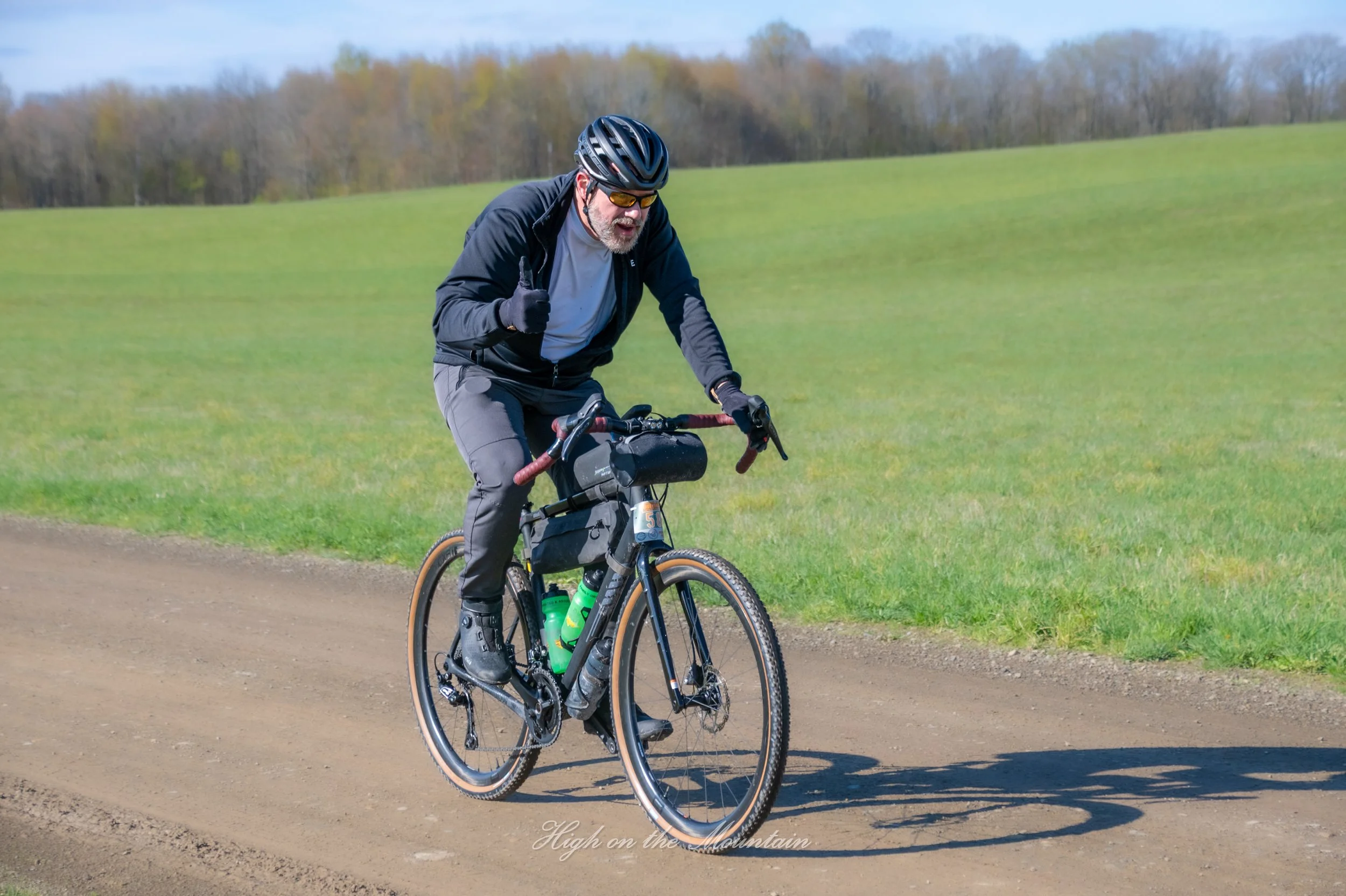 A man with a beard and glasses wearing a black helmet, black jacket, and gray pants riding a black mountain bike with green water bottles on a dirt trail through a grassy field with trees in the background, giving a thumbs up.