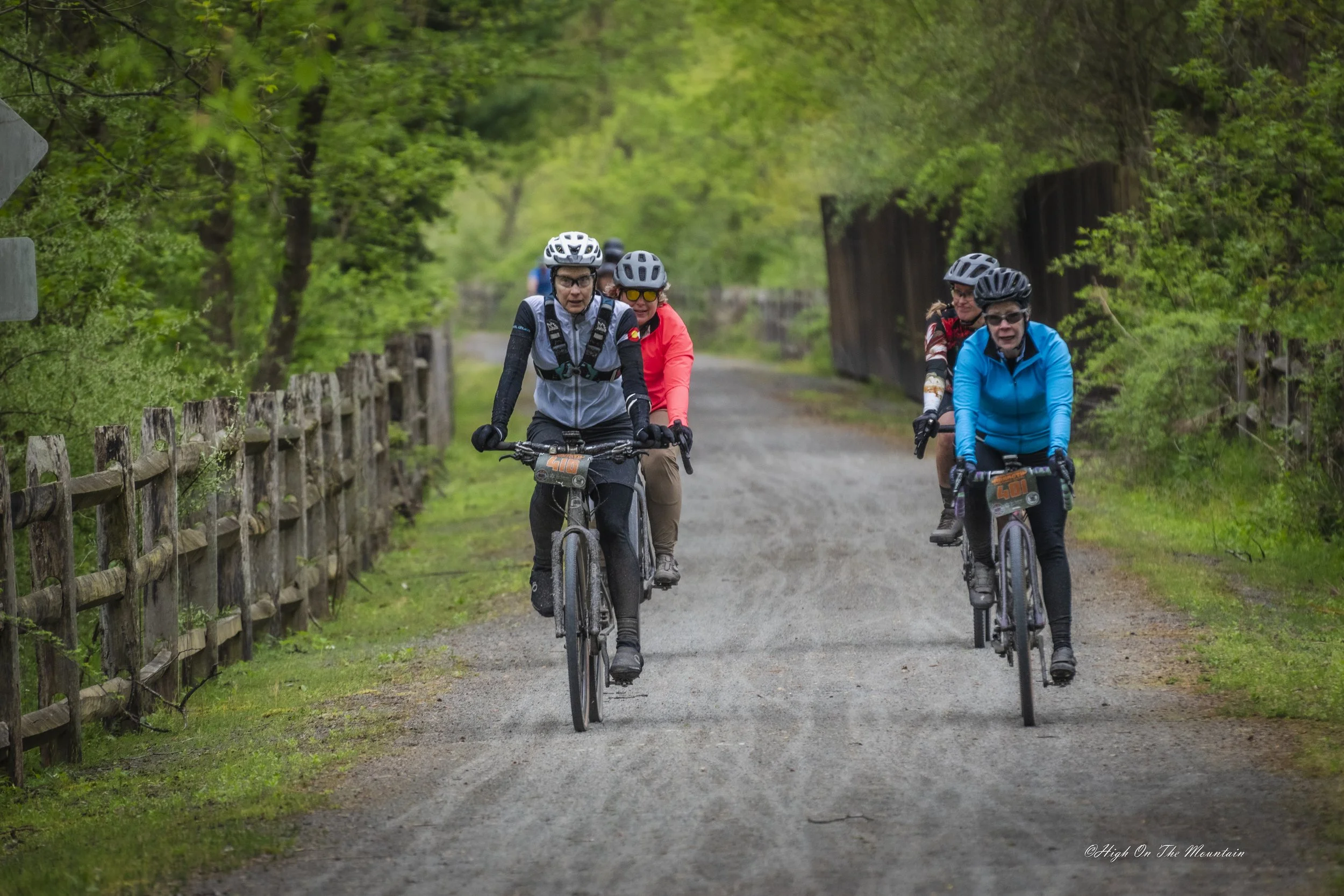 Group of four women riding mountain bikes on a gravel trail through a lush green forest, wearing helmets and colorful jackets.