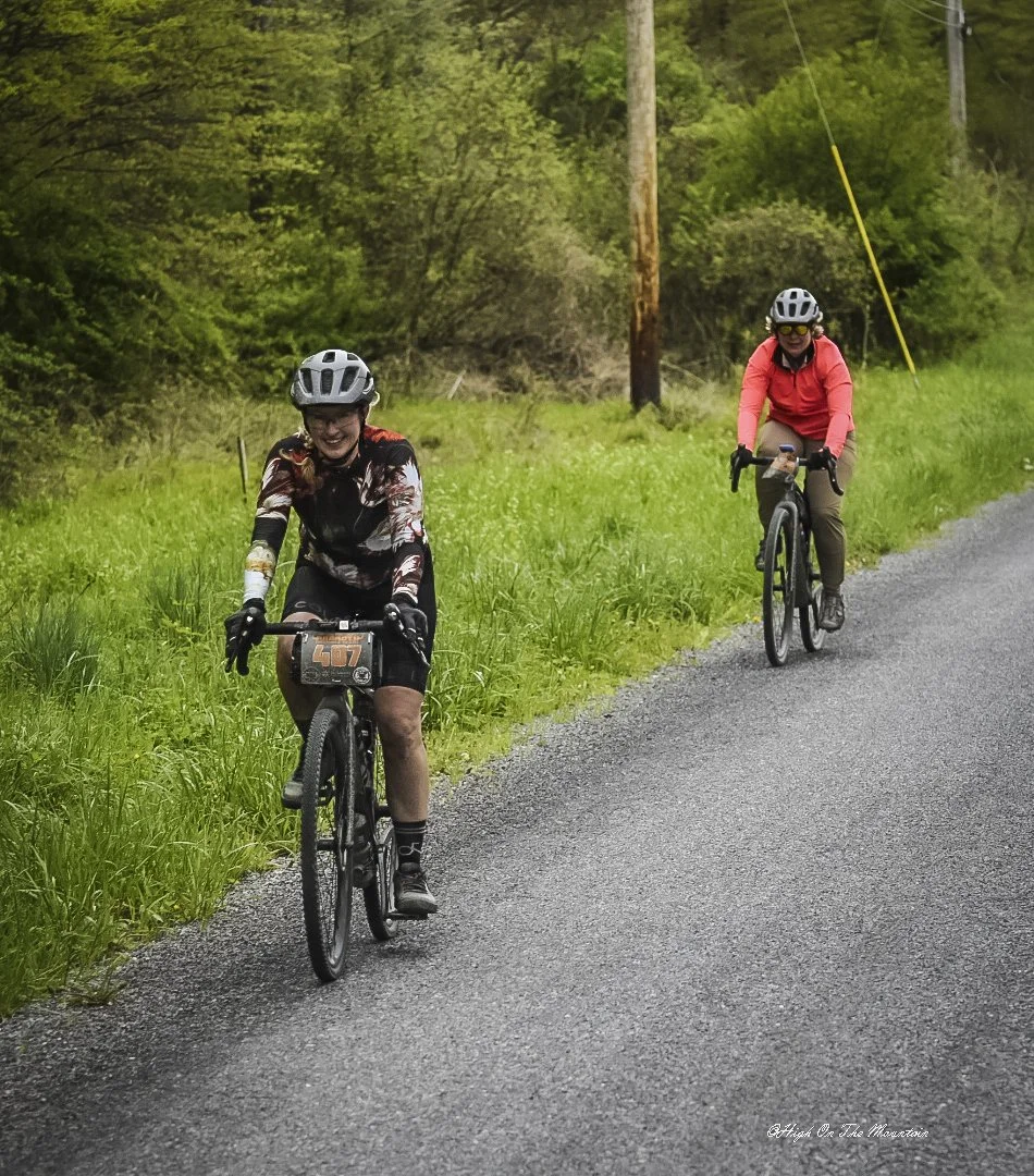 Two women riding mountain bikes on a gravel trail through a lush green forest, wearing helmets and outdoor clothing.