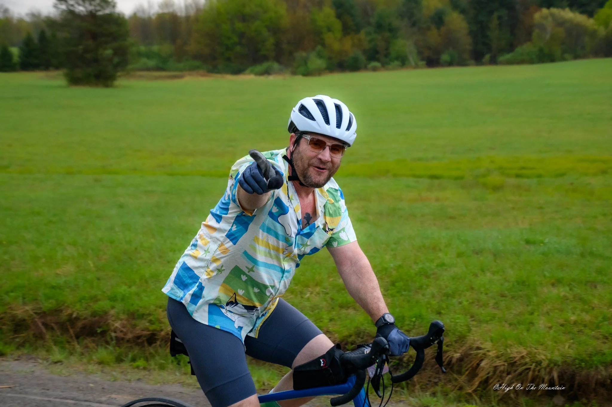 A man biking outdoors on a grassy trail with trees in the background, wearing a white bike helmet, glasses, a colorful patterned shirt, and dark shorts, smiling and pointing at the camera.