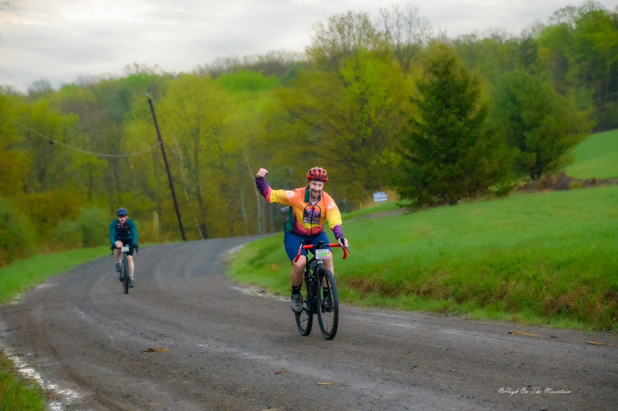 Two cyclists riding on a gravel country road surrounded by green trees and grassy fields, with one cyclist in the foreground raising his fist and smiling.