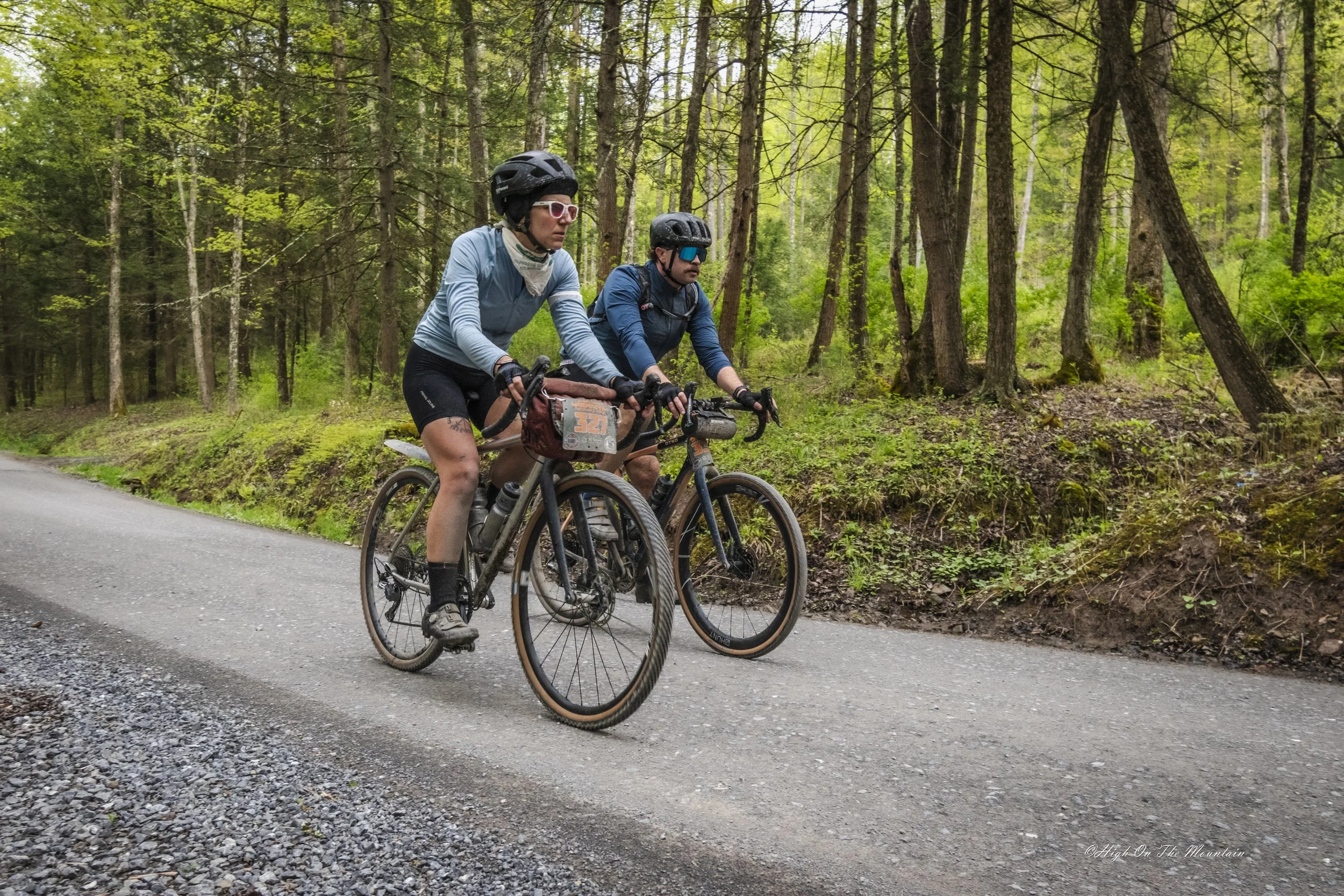 Two cyclists riding mountain bikes on a gravel trail through a forest with green trees.