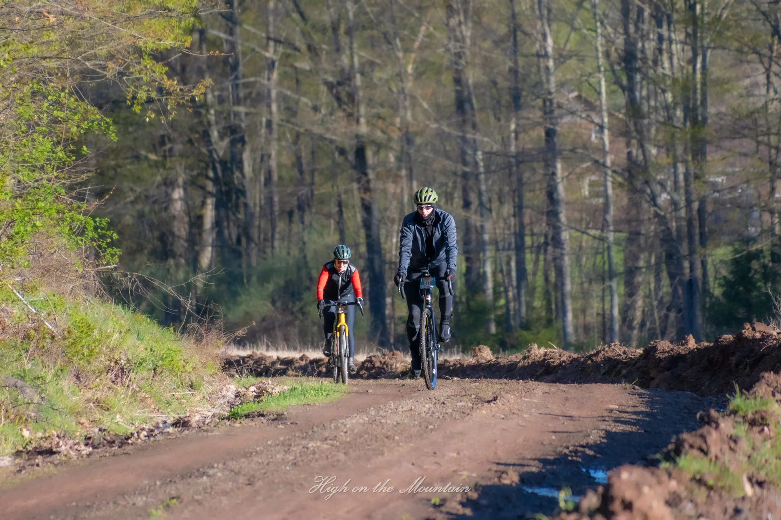 Two cyclists riding mountain bikes along a dirt trail through a wooded area with tall trees and mostly green leaves, under a partly cloudy sky.