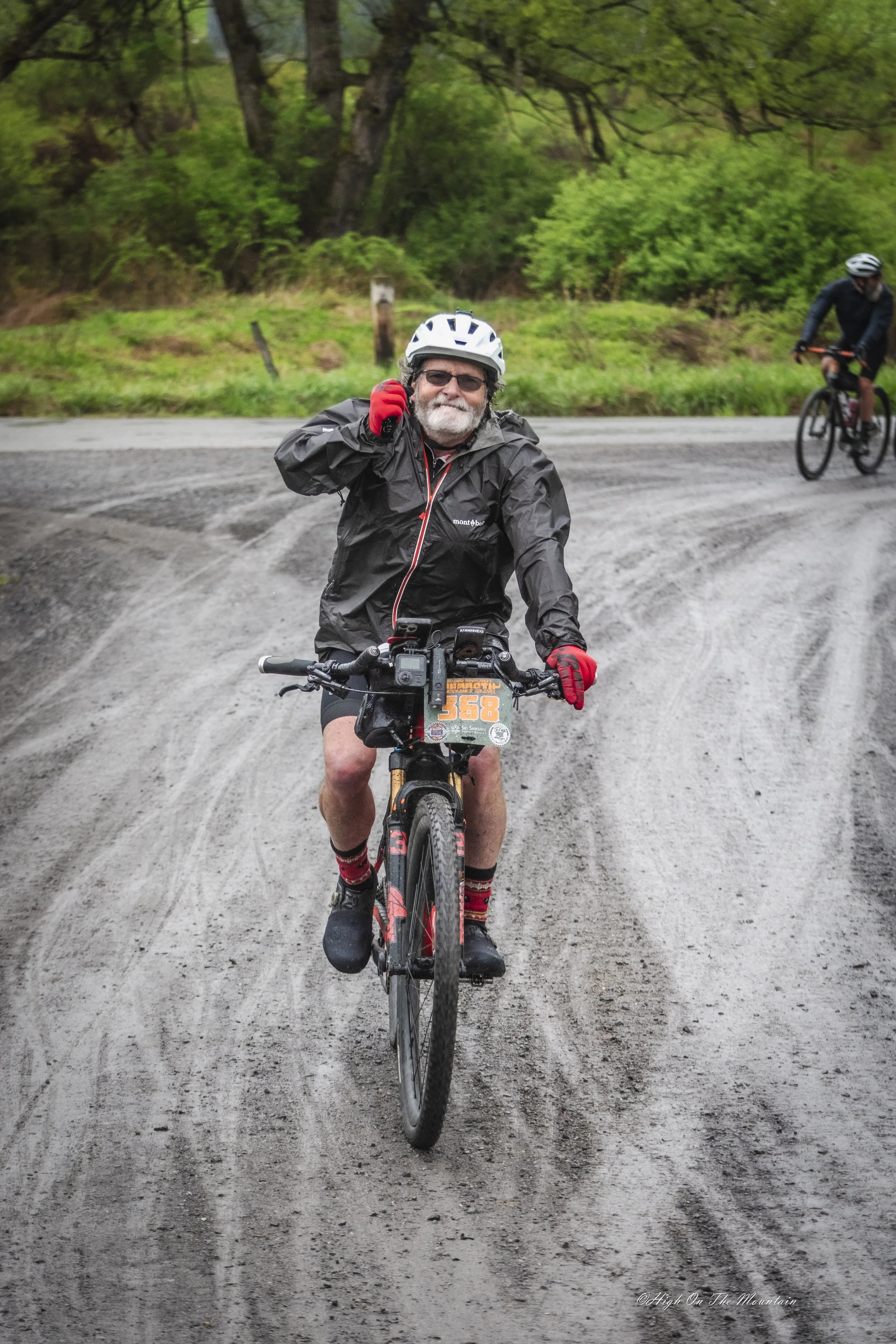 An older man with gray hair and beard riding a mountain bike on a dirt trail, wearing a white helmet, black jacket, red gloves, and red and black socks. In the background, another cyclist rides on a similar trail surrounded by green trees and grass.