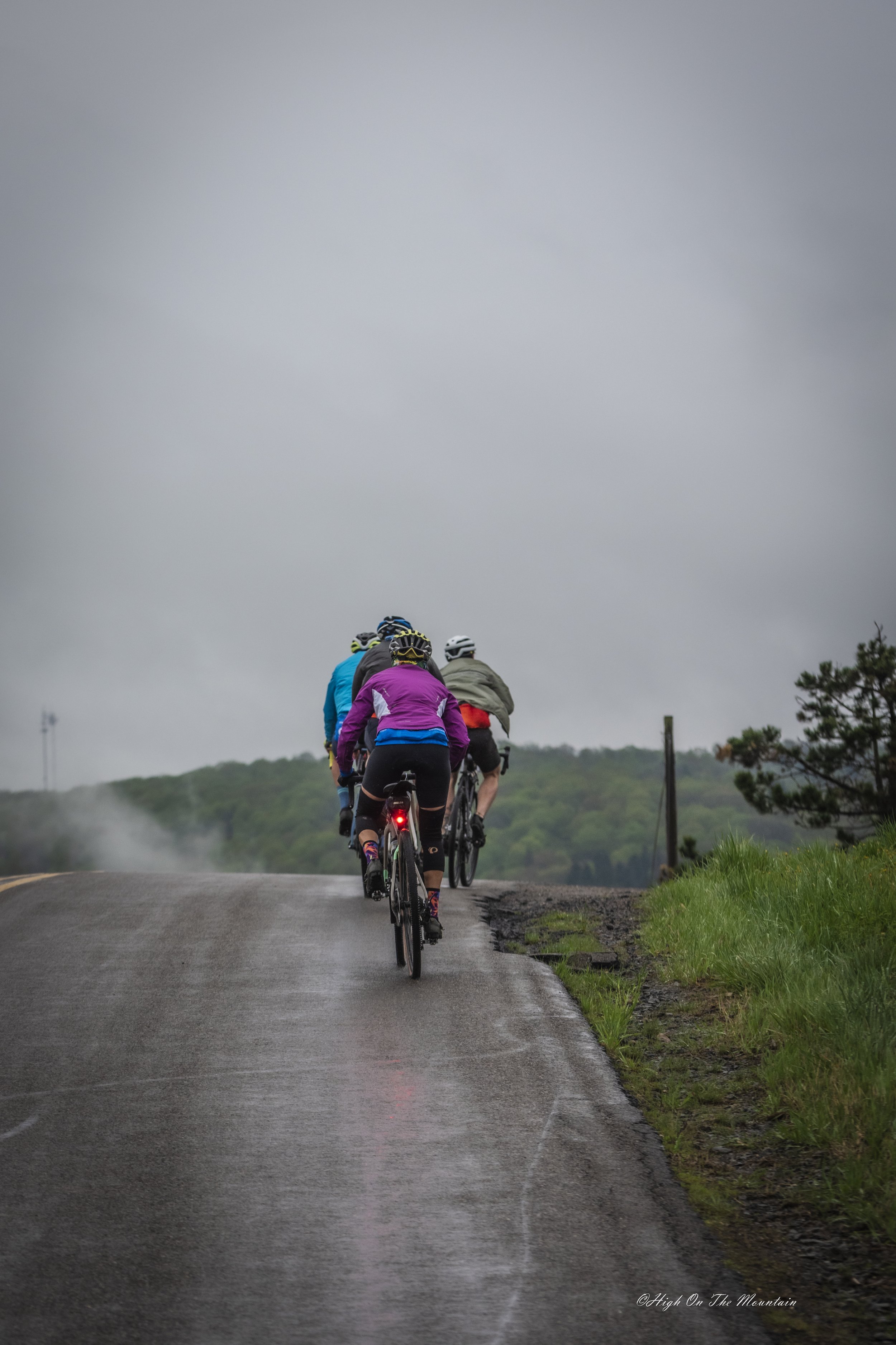 A group of cyclists riding on a wet, paved road through a green countryside landscape, under a cloudy sky.