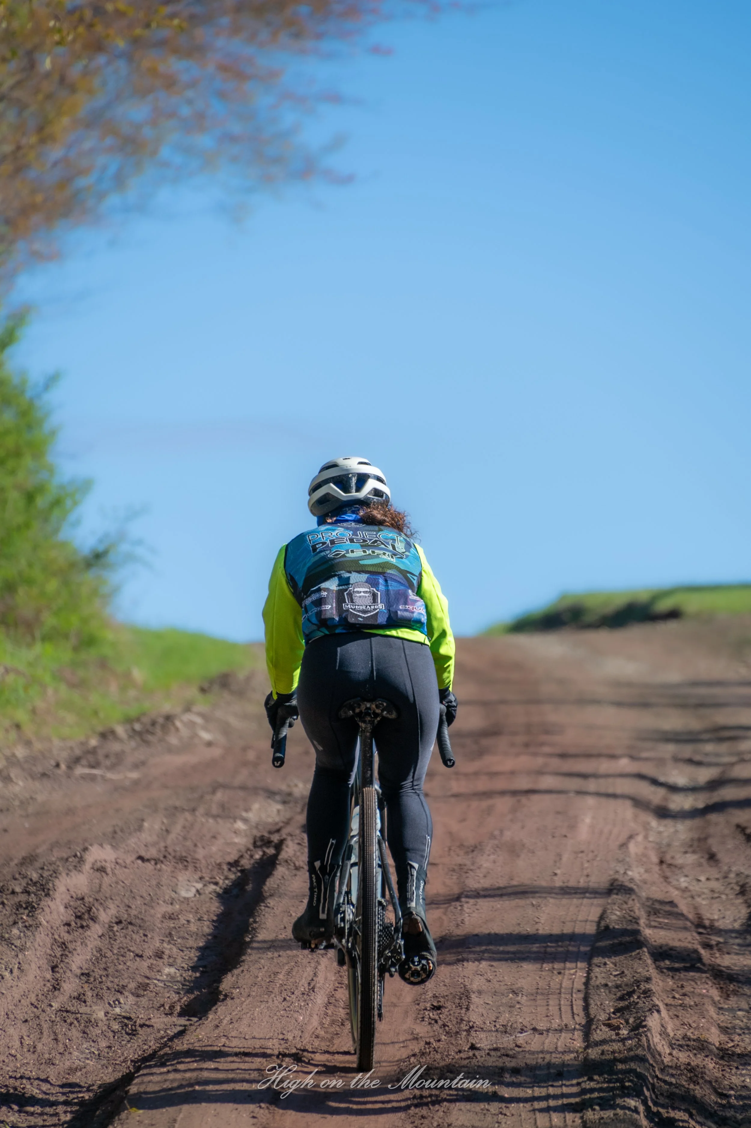 A person wearing a helmet and cycling attire riding a mountain bike on a dirt trail, with grassy hills and a clear blue sky in the background.