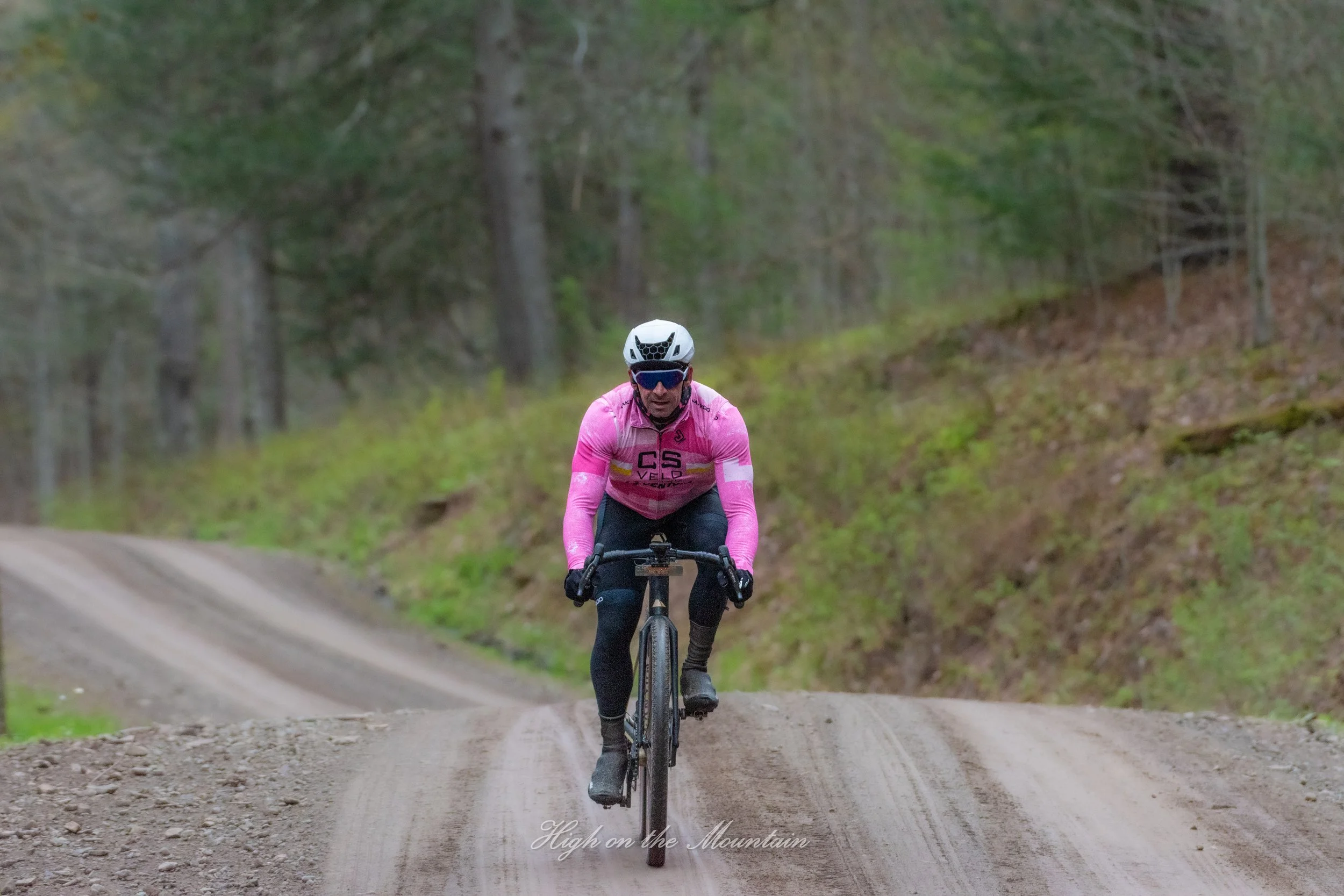 A mountain biker wearing a pink jersey, helmet, and sunglasses rides on a dirt trail through a forest with green trees.